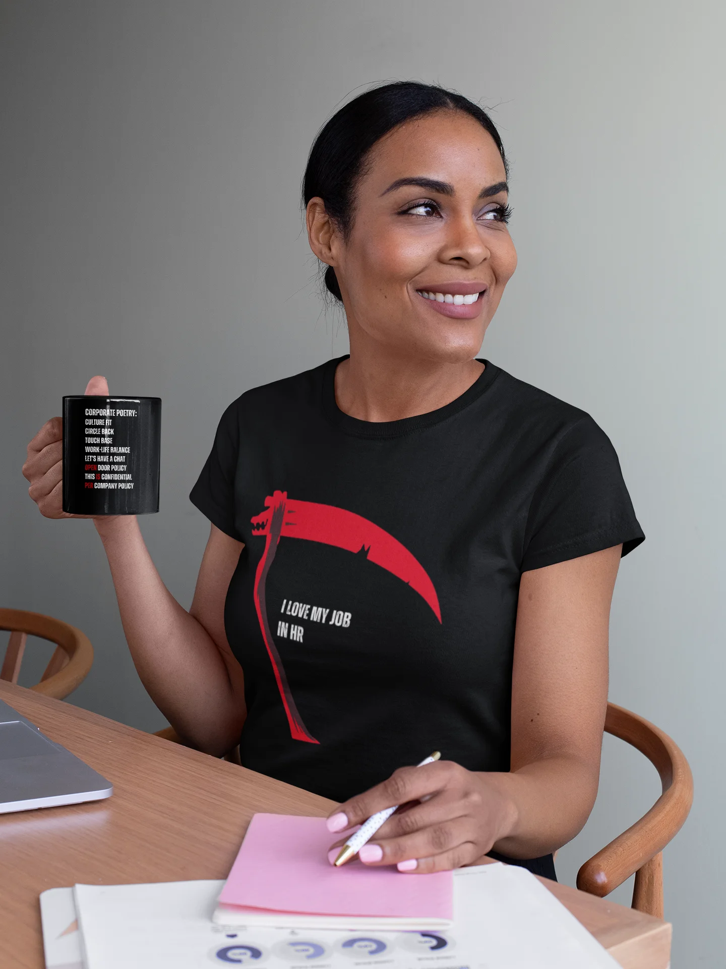 Woman holding a mug and wearing a black t-shirt with a red graphic design, sitting at a table.