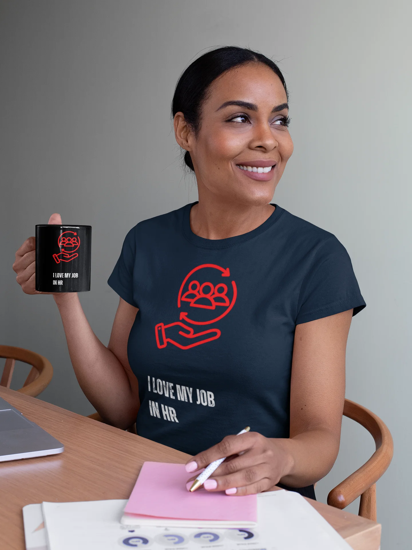  I love my Job T-Shirt Woman holding a mug with a design and text, wearing a dark t-shirt with a red graphic and text, sitting at a desk.