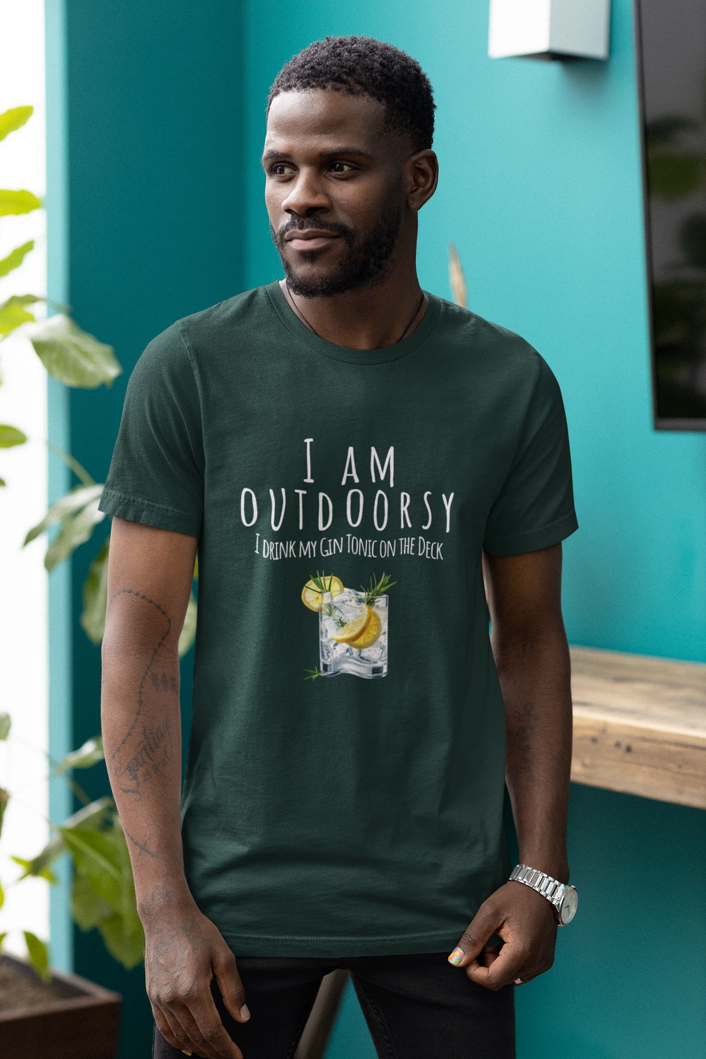 Man wearing a green t-shirt with 'I AM OUTDOORSY' text and graphic, standing against a teal wall.