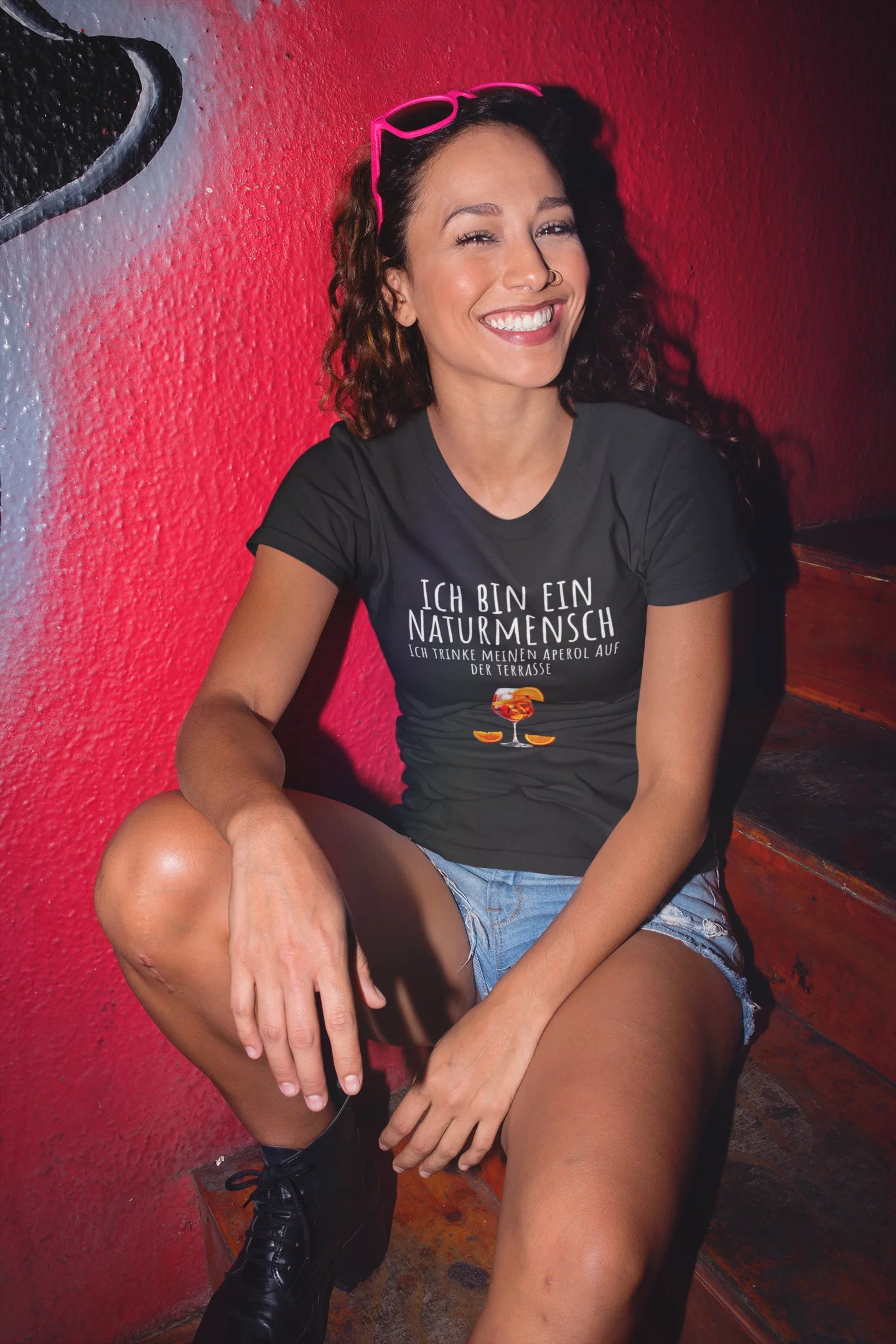 Woman sitting on a red wall wearing a black t-shirt with text and graphics.
