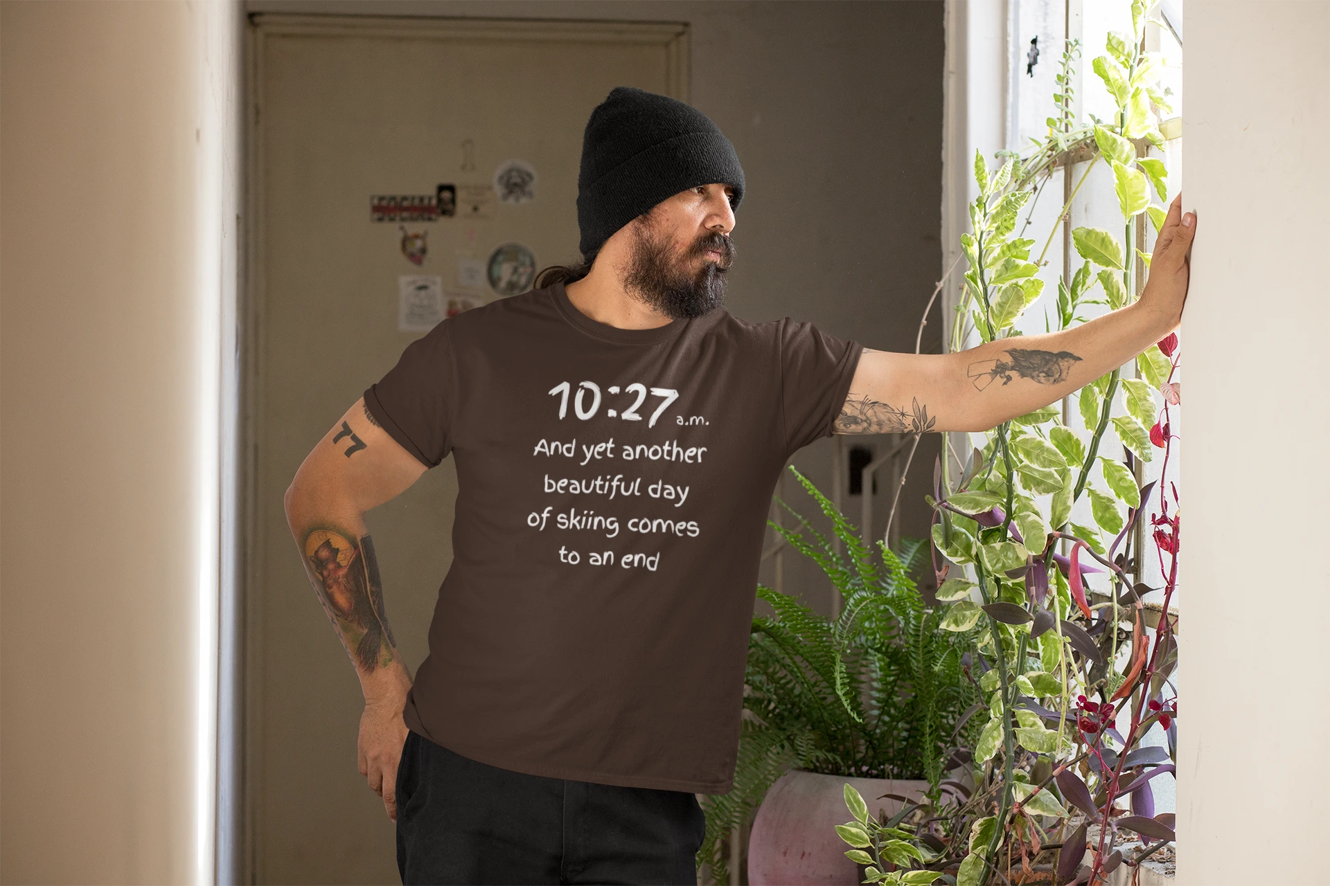 Man wearing a brown t-shirt with text, standing indoors next to plants.