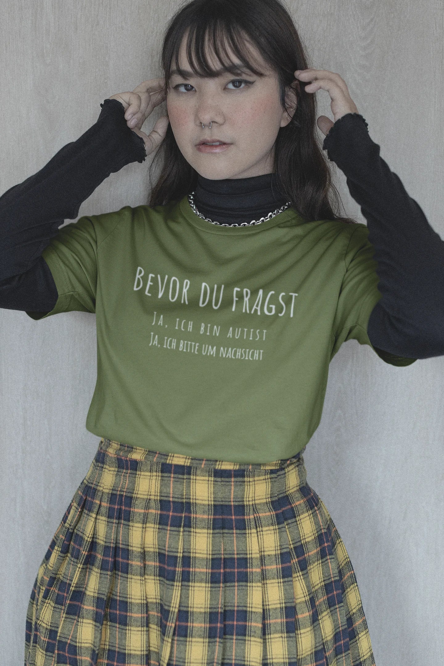 Person wearing a green t-shirt with text and a plaid skirt against a plain background