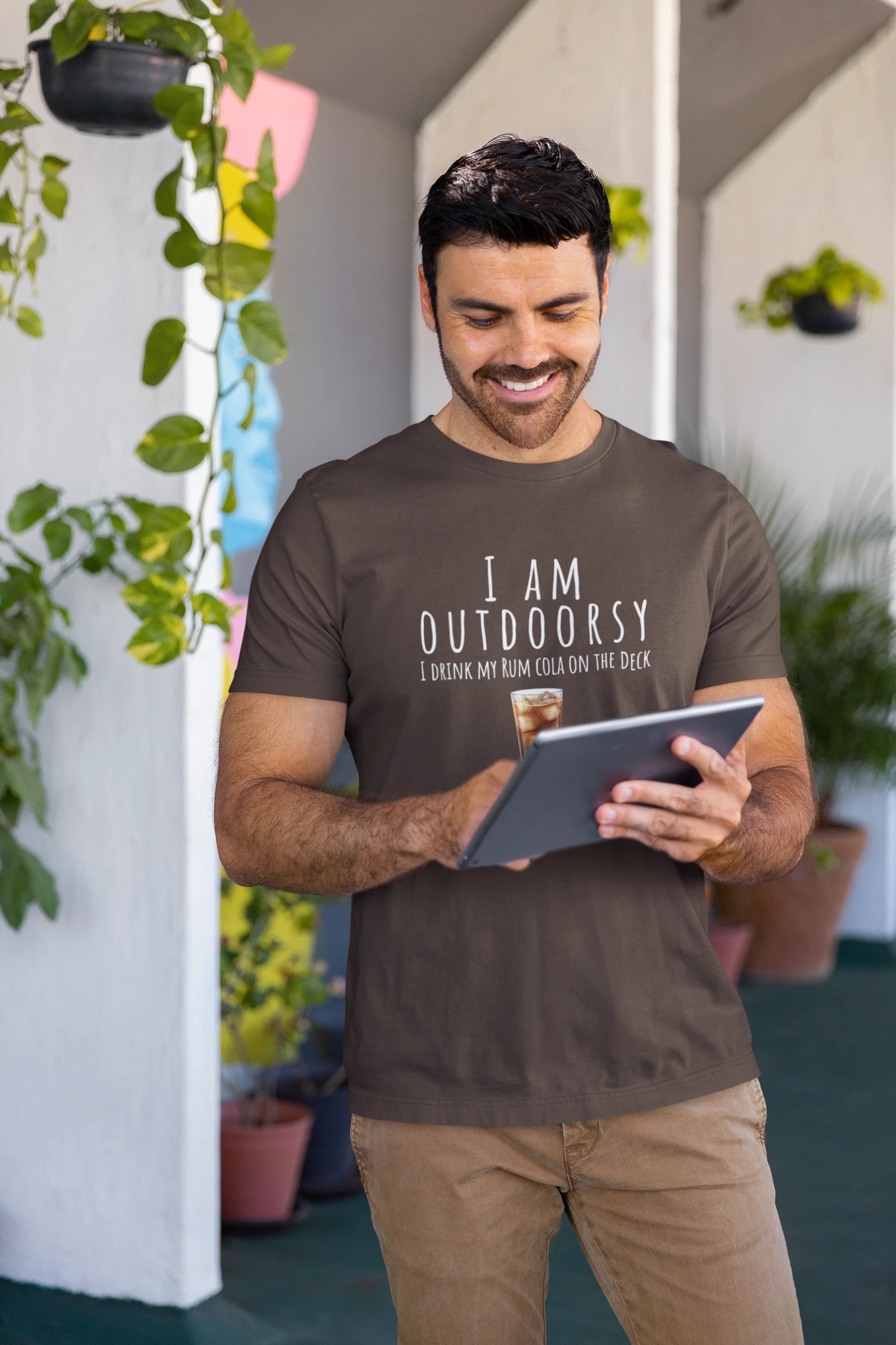 Man wearing a 'I AM OUTDOORSY' t-shirt, holding a tablet, surrounded by plants.