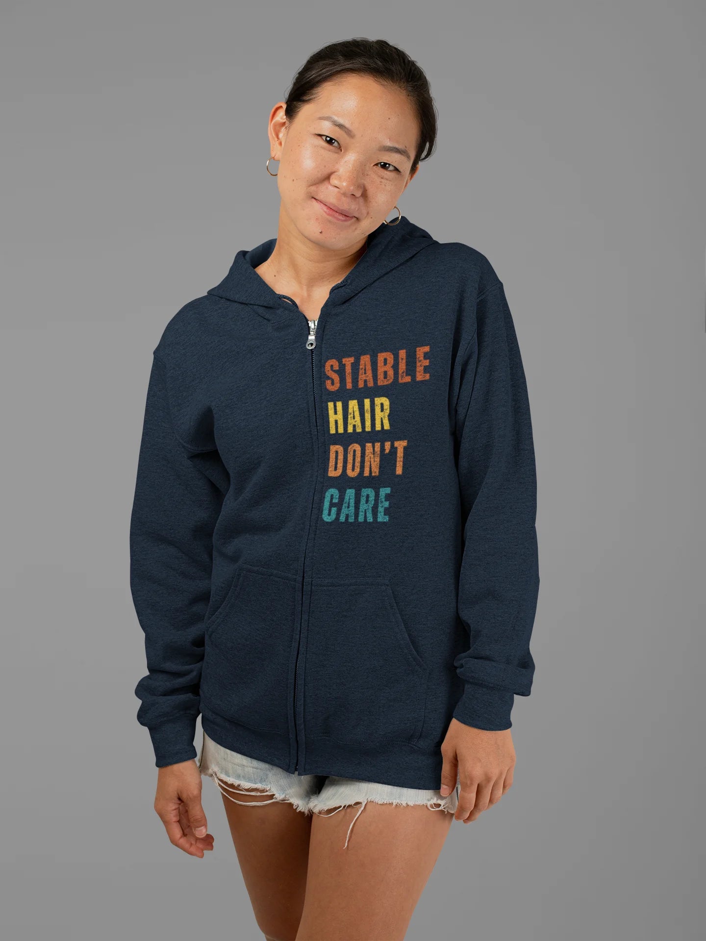 Equestrian Hoodie with Stable Hair Don't Care text and horse barn back print. Person wearing a navy blue hoodie with colorful text on a gray background
