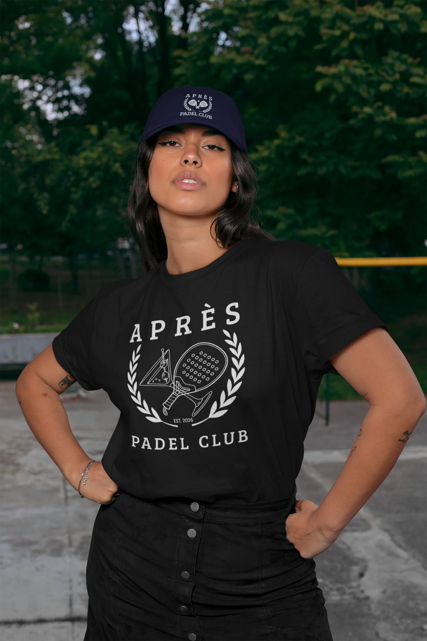 Padel Cap Woman wearing a black t-shirt with 'Ap
es Padel Club' design and a cap, standing outdoors.