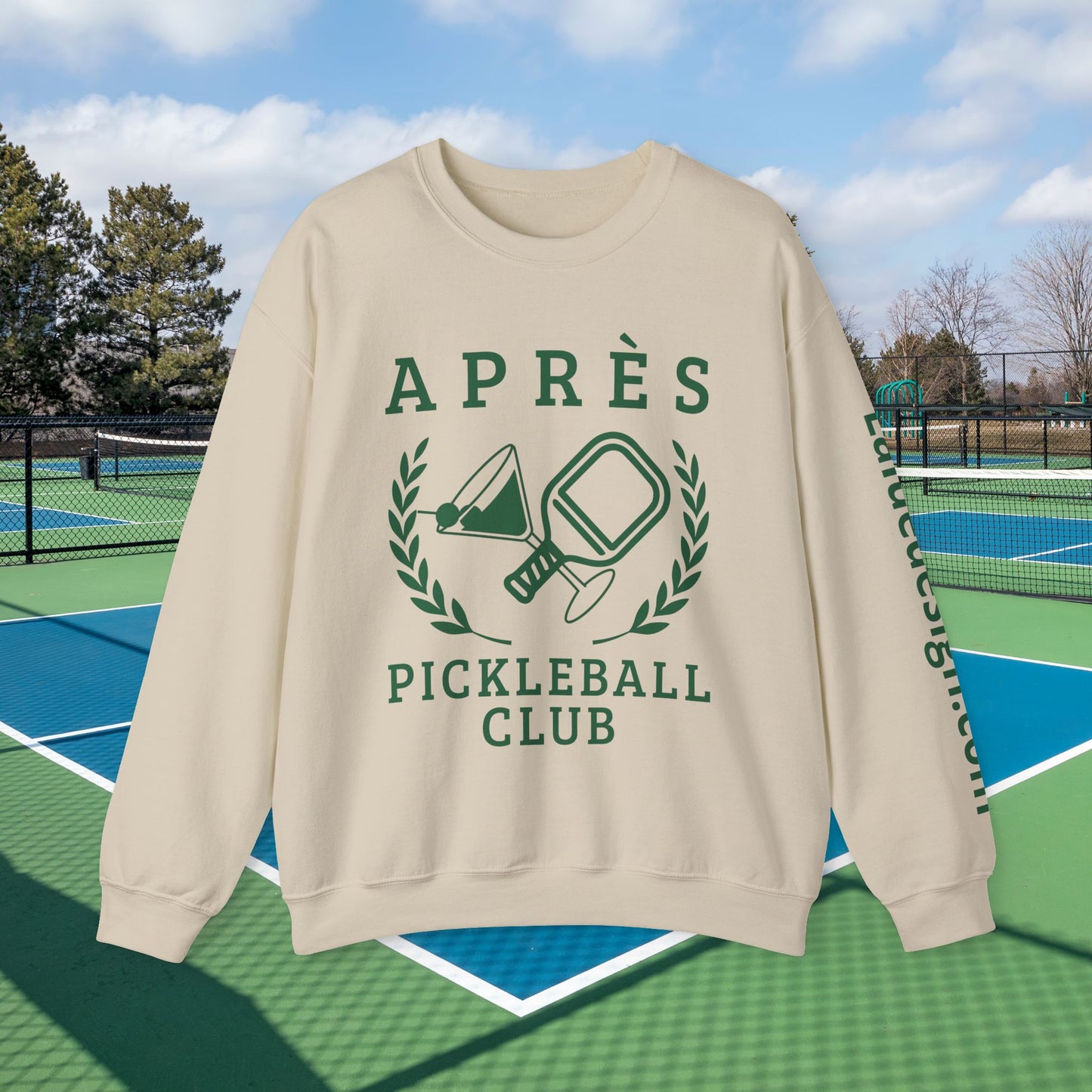 personalized padel gift A beige crewneck sweatshirt with a green graphic print reading 'APRÈS PICKLEBALL CLUB' and featuring pickleball-themed illustrations, shown on a green and blue outdoor pickleball court.