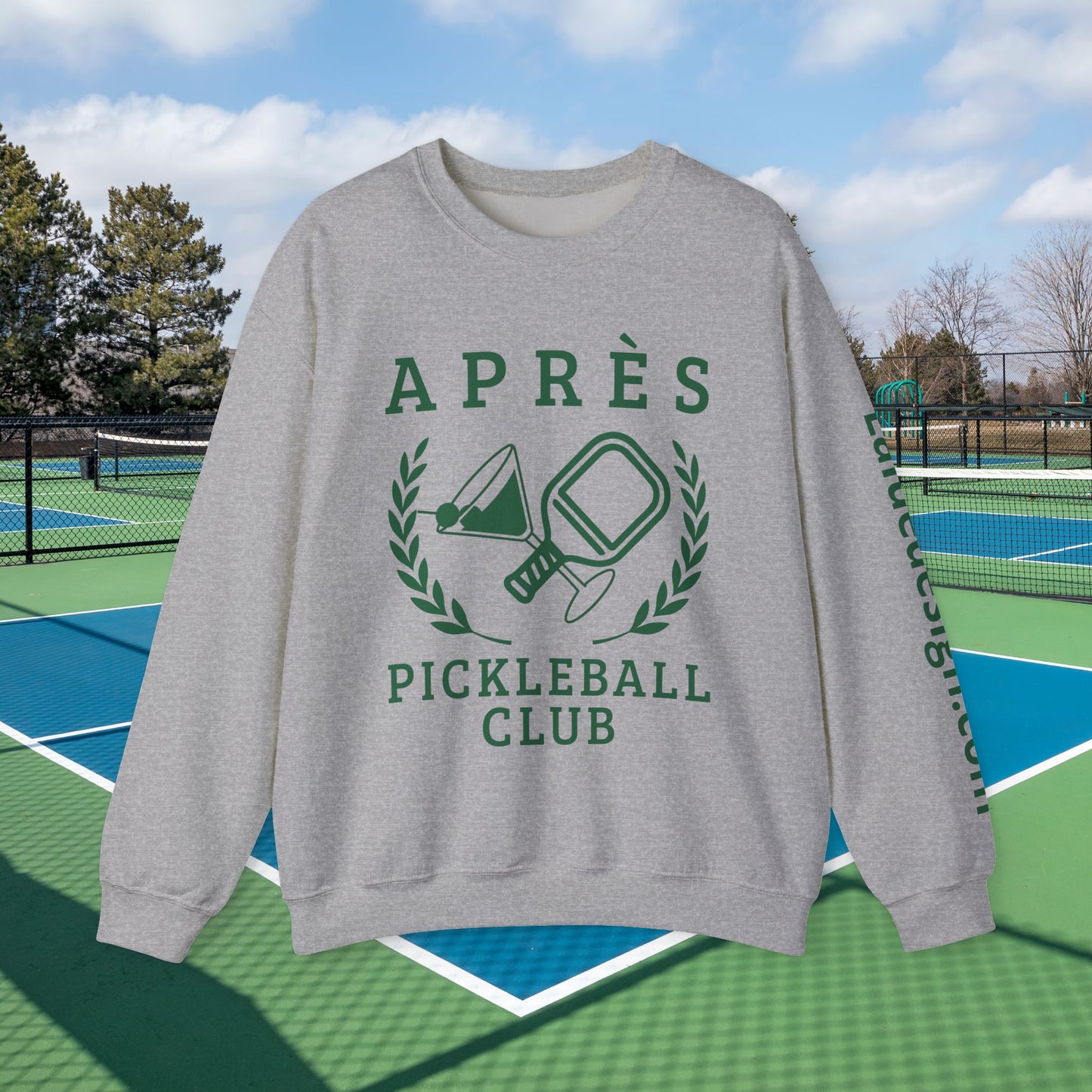 personalized padel gift A light gray crewneck sweatshirt with a green graphic print reading 'APRÈS PICKLEBALL CLUB' and showing pickleball rackets and a paddle, displayed against a background of outdoor pickleball courts under a blue sky.