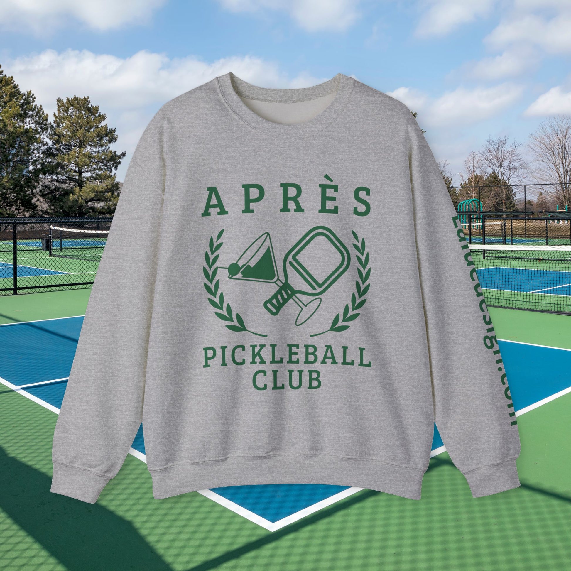 personalized padel gift A light gray crewneck sweatshirt with a green graphic print reading 'APRÈS PICKLEBALL CLUB' and showing pickleball rackets and a paddle, displayed against a background of outdoor pickleball courts under a blue sky.