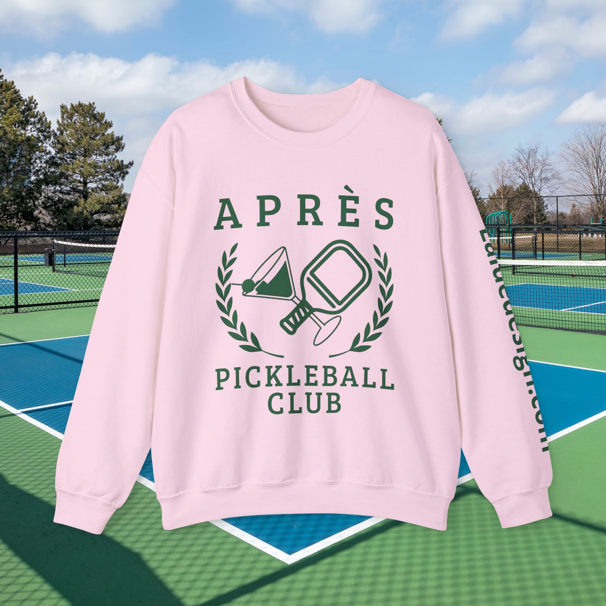 personalized padel gift A light pink crewneck sweatshirt with a dark green pickleball-themed graphic reading 'APRÈS PICKLEBALL CLUB' on the front, displayed against an outdoor pickleball court background.