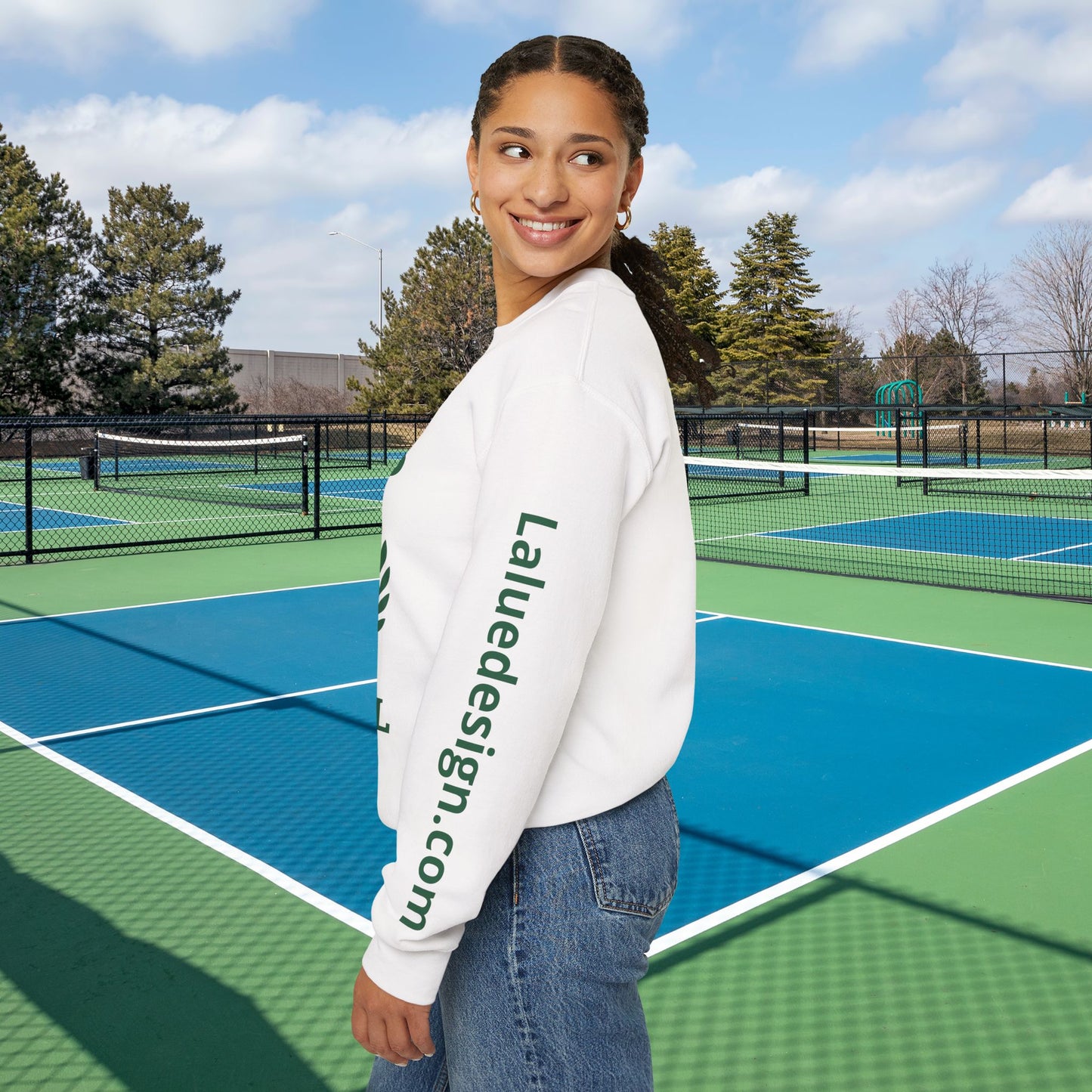 A woman stands on an outdoor tennis court wearing a white crewneck sweatshirt with green text reading 'Laluedesign.com' printed down the left sleeve, paired with blue jeans.