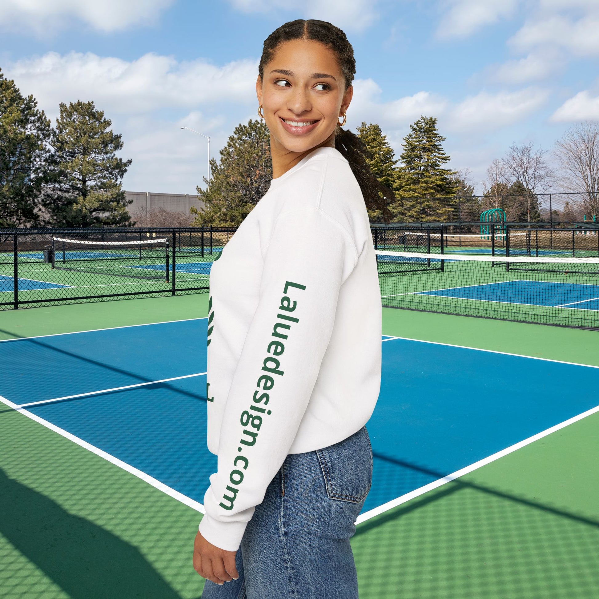 A woman stands on an outdoor tennis court wearing a white crewneck sweatshirt with green text reading 'Laluedesign.com' printed down the left sleeve, paired with blue jeans.