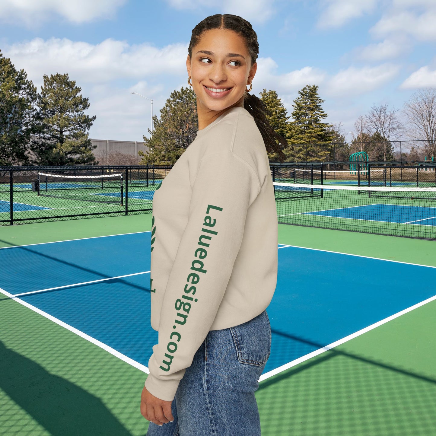 personalized padel gift A woman stands on a blue and green tennis court wearing a beige crewneck sweatshirt with 'Laluedesign.com' printed in dark green down the left sleeve, paired with blue jeans.
