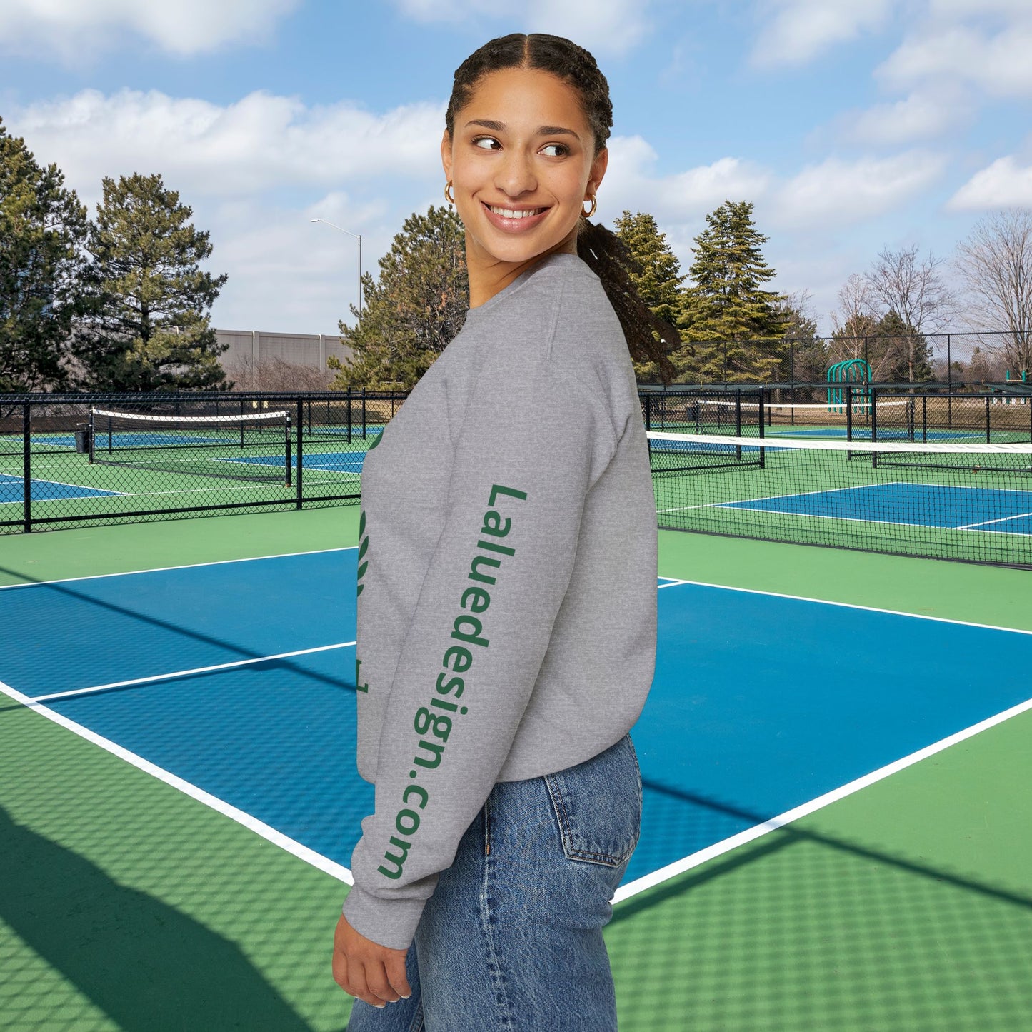 personalized padel gift A woman stands on a blue and green outdoor tennis court wearing a heather gray sweatshirt with green text reading 'Laluedesign.com' down the sleeve, paired with blue jeans.