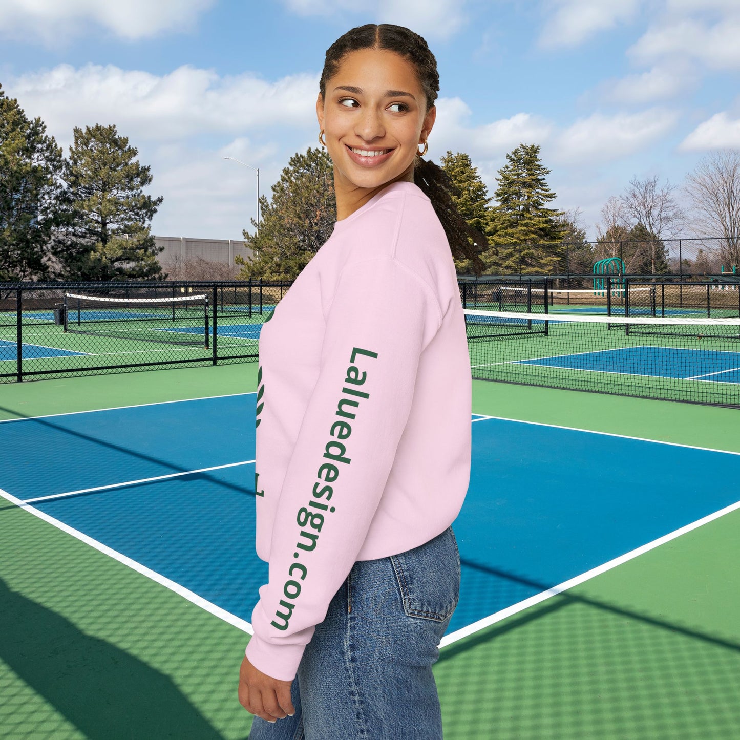 personalized padel gift A woman stands on an outdoor tennis court wearing a light pink crewneck sweatshirt with 'Laluedesign.com' printed vertically in dark green on the left sleeve, paired with blue jeans.