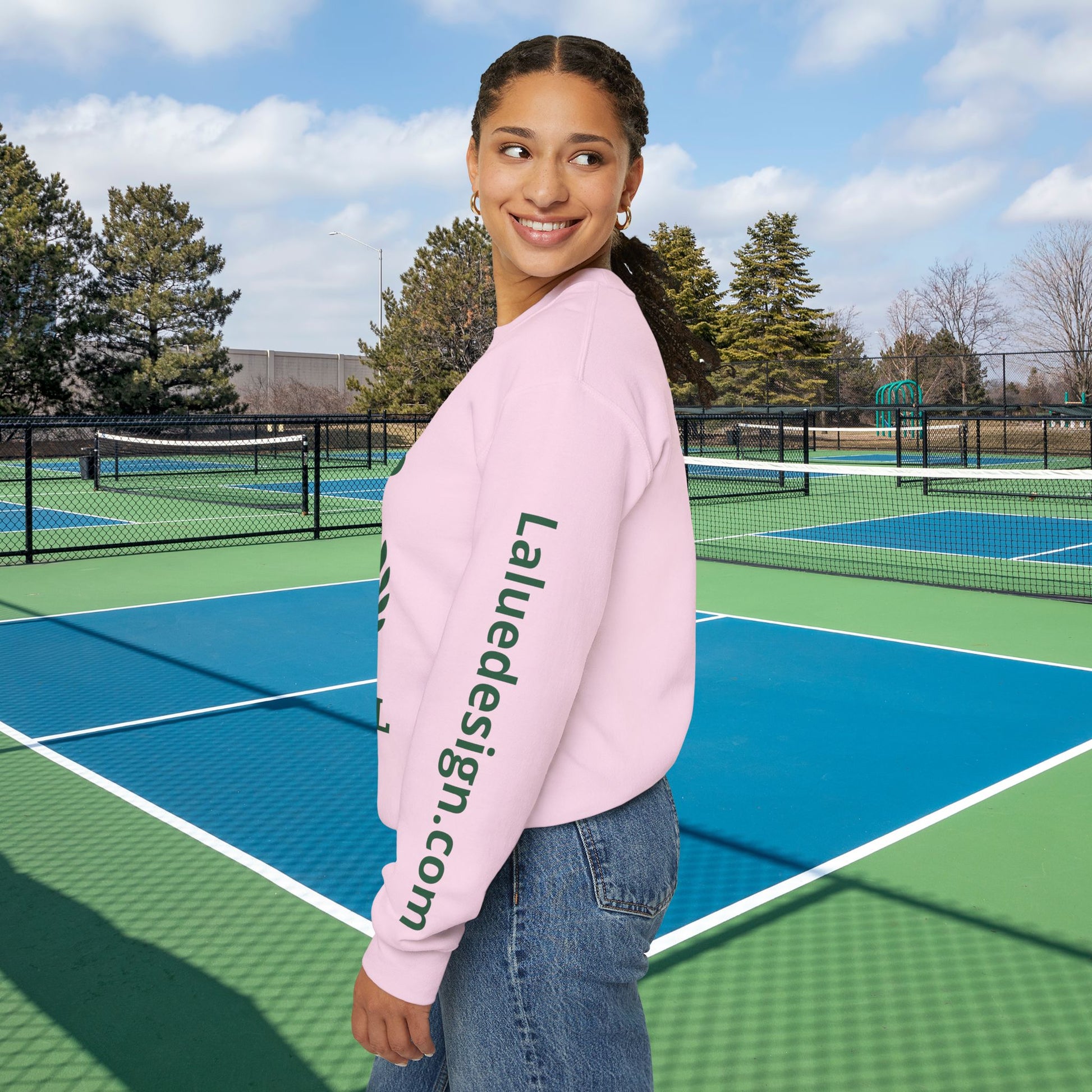 personalized padel gift A woman stands on an outdoor tennis court wearing a light pink crewneck sweatshirt with 'Laluedesign.com' printed vertically in dark green on the left sleeve, paired with blue jeans.