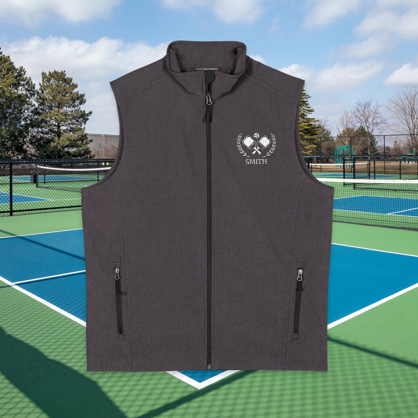 personalized pickleball gift A dark gray sleeveless zip-front vest with two vertical zip pockets and a white chest graphic featuring crossed tennis rackets, laurel branches, and the name 'SMITH', shown against a tennis court background.