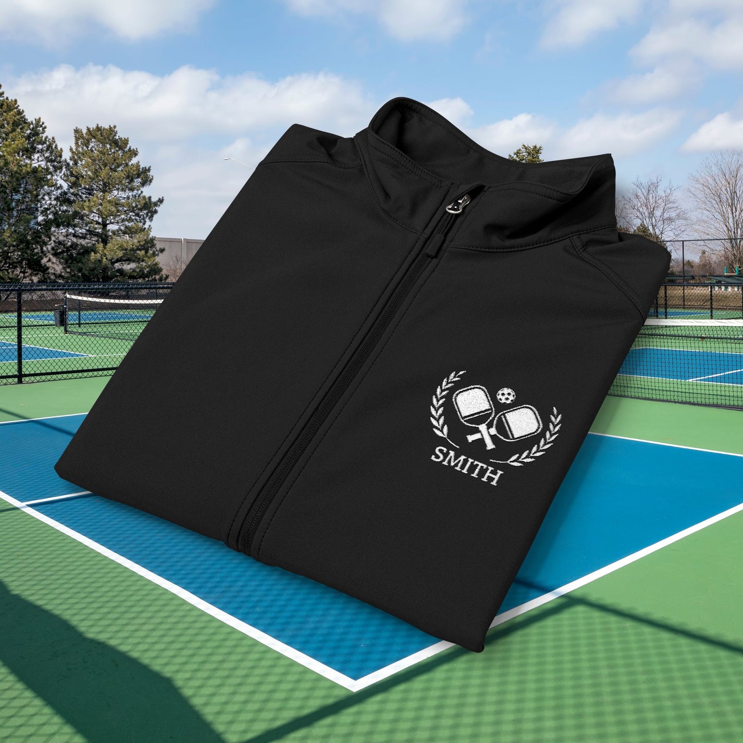personalized pickleball gift A black zip-up jacket with a white embroidered crest featuring crossed tennis rackets, a soccer ball, and the name 'SMITH' below, displayed on a blue and green tennis court with trees and sky in the background.