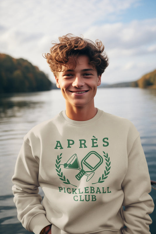 personalized padel gift A smiling young man with curly brown hair stands outdoors by a lake wearing a cream sweatshirt with a green 'APRÈS PICKLEBALL CLUB' graphic on the front.