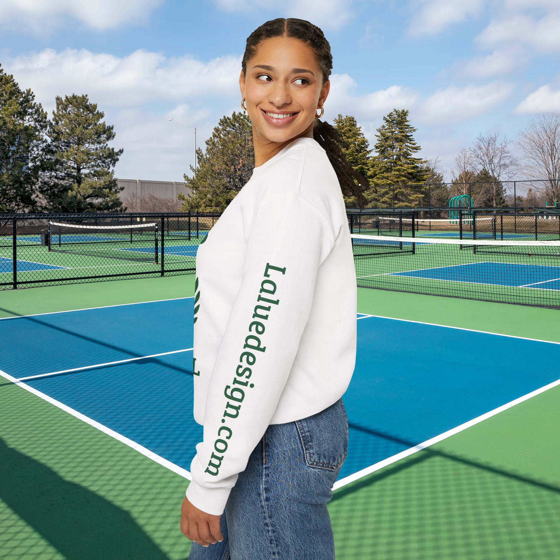 personalized padel gift A woman stands on an outdoor tennis court wearing a white crewneck sweatshirt with green text reading 'Laluedesign.com' printed down the left sleeve, paired with blue jeans.