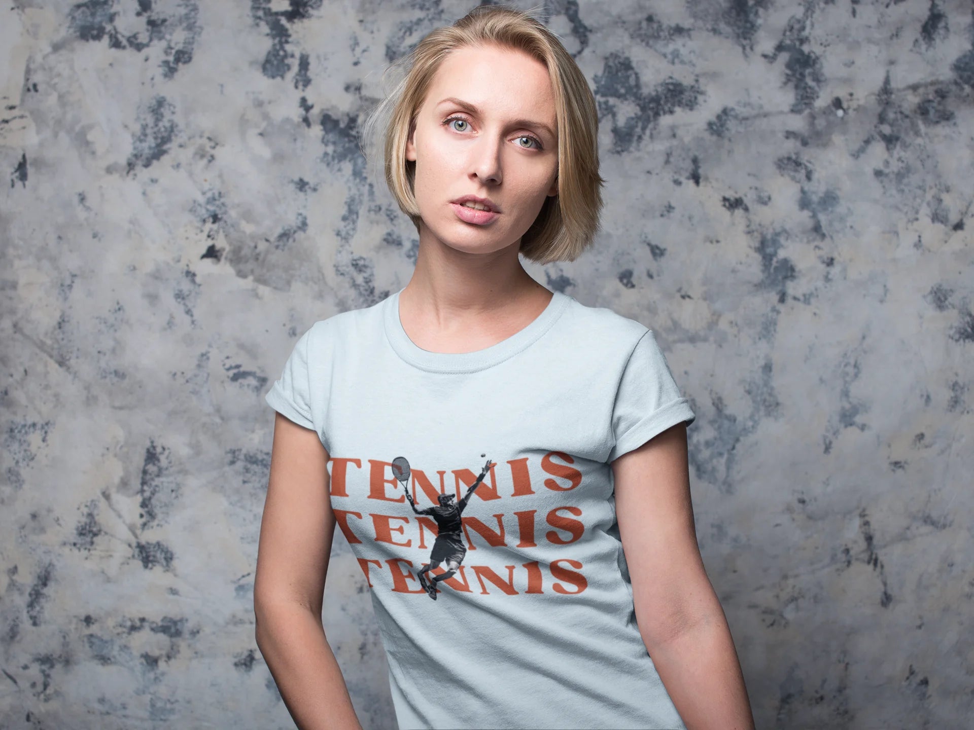 Woman wearing a light blue t-shirt with 'TENNIS' printed on it against a textured gray background