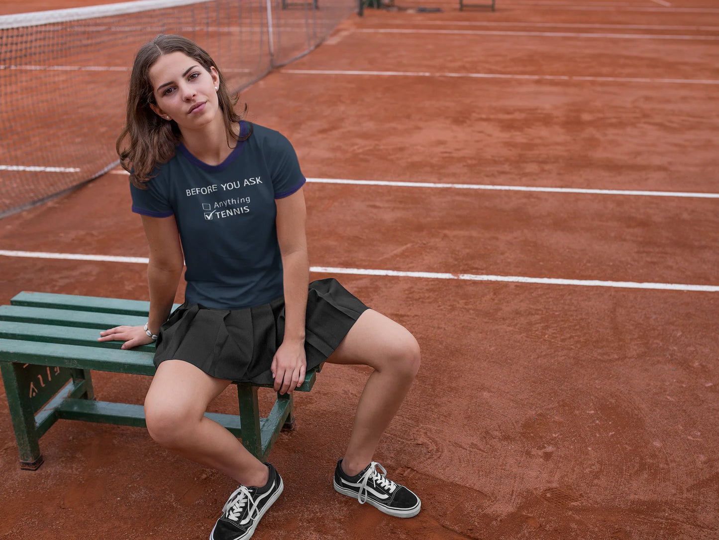 Funny Tennis T-Shirt Woman sitting on a bench on a clay tennis court wearing a navy blue t-shirt and skirt.