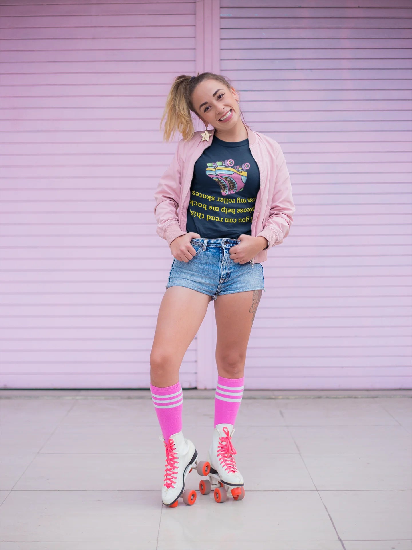 Roller skate shirt A woman posing on roller skates in front of a pink metal shutter wall. She is wearing a black graphic t-shirt, a light pink cardigan, denim shorts, bright pink knee-high socks, and white roller skates with pink laces and orange wheels.