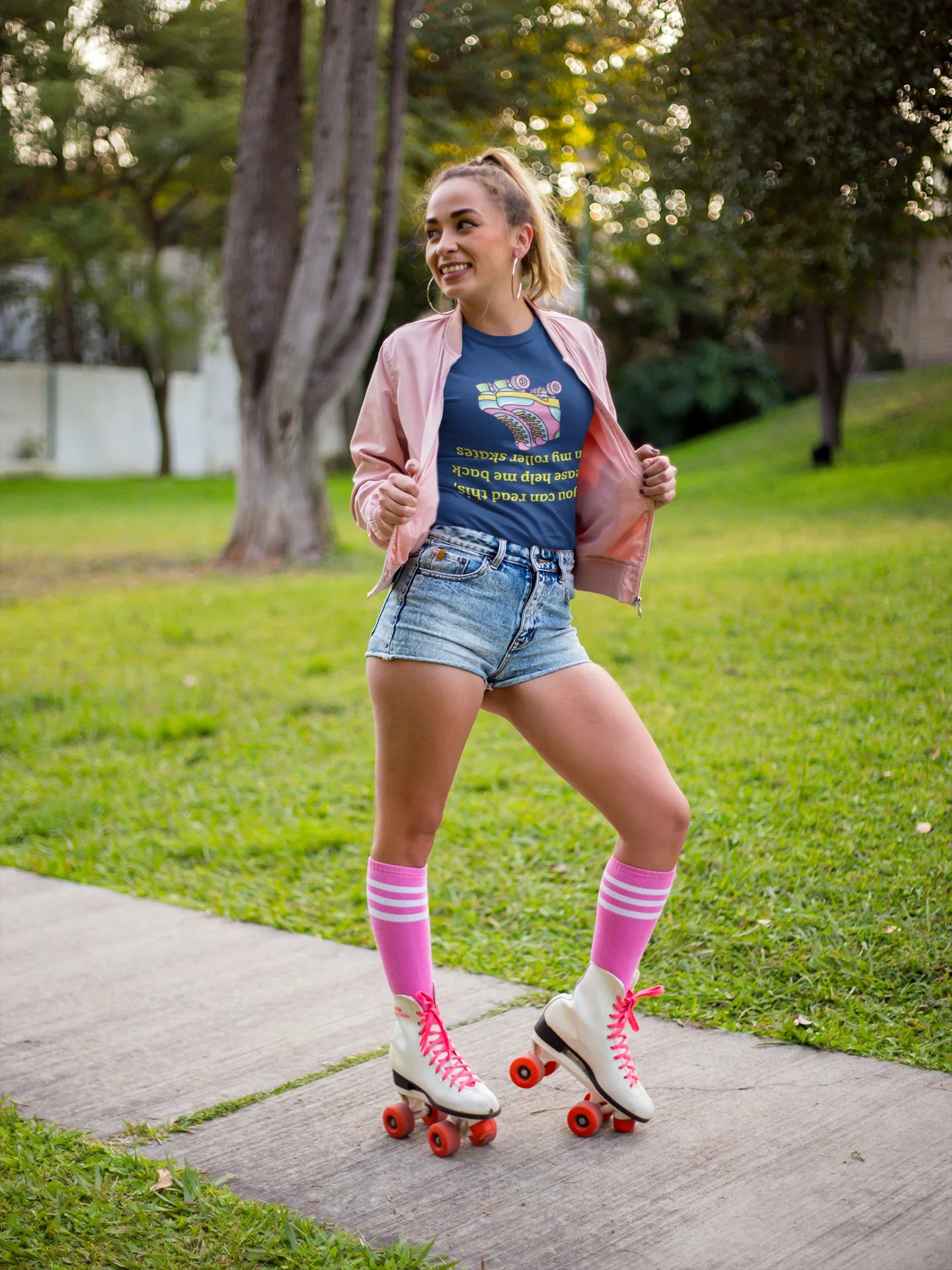 Roller skate shirt A woman wearing a pink jacket, blue graphic t-shirt, denim shorts, pink knee-high socks, and white roller skates with red wheels poses on a concrete path in a grassy park.