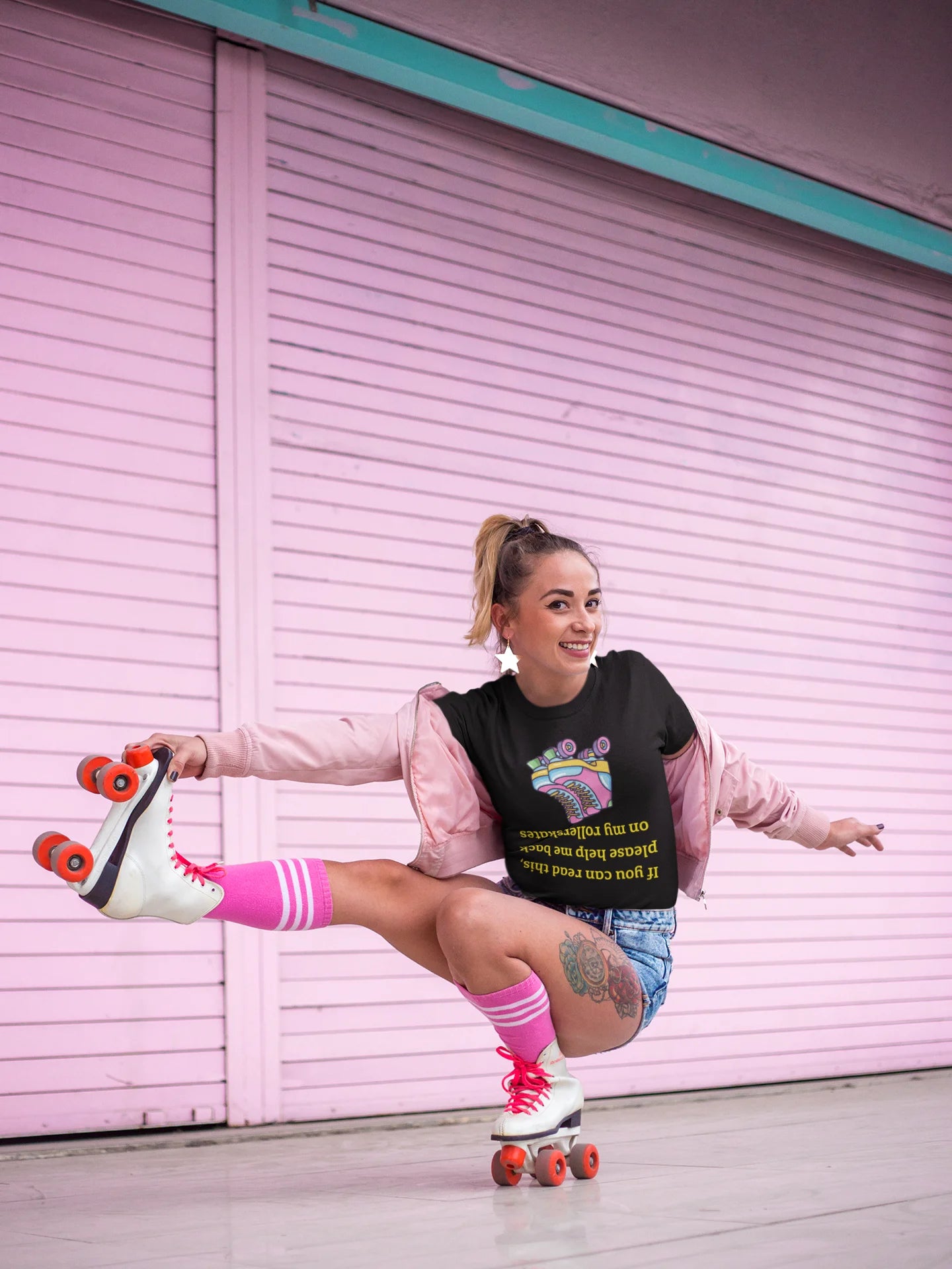 Roller skate shirt A woman posing on a white quad roller skate in front of a pink metal garage door. She is wearing a black graphic t-shirt, a light pink zip-up jacket, denim shorts, and pink knee-high socks.