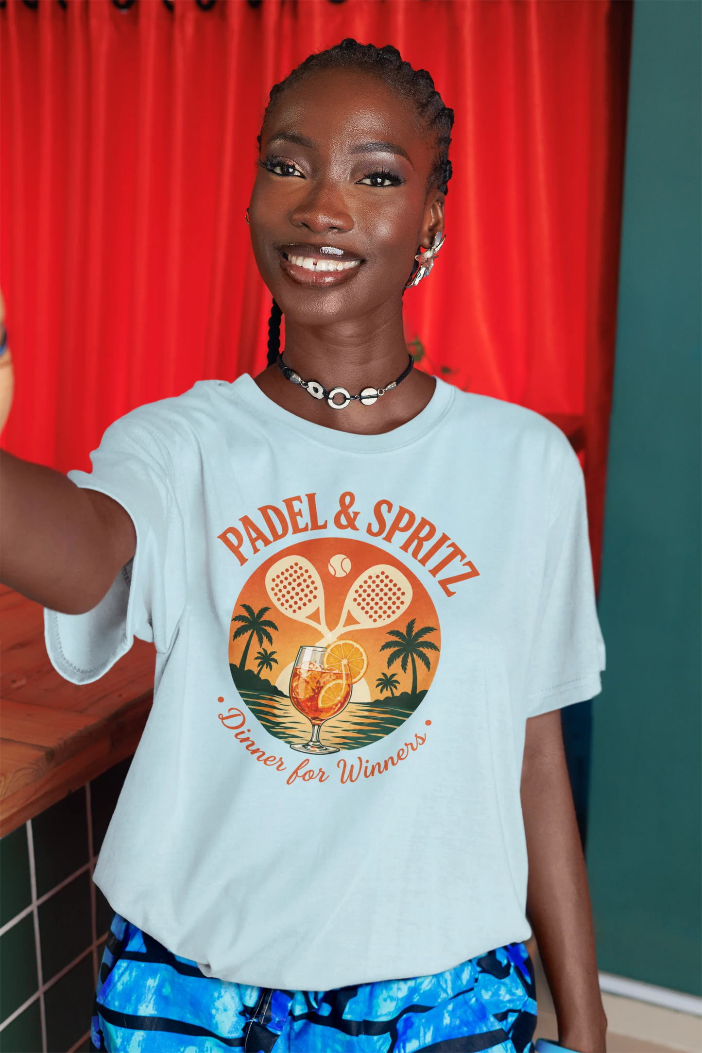 Padel Aperol T-Shirt Person wearing a light blue t-shirt with 'Padel & Spritz' design, standing in front of a red curtain.