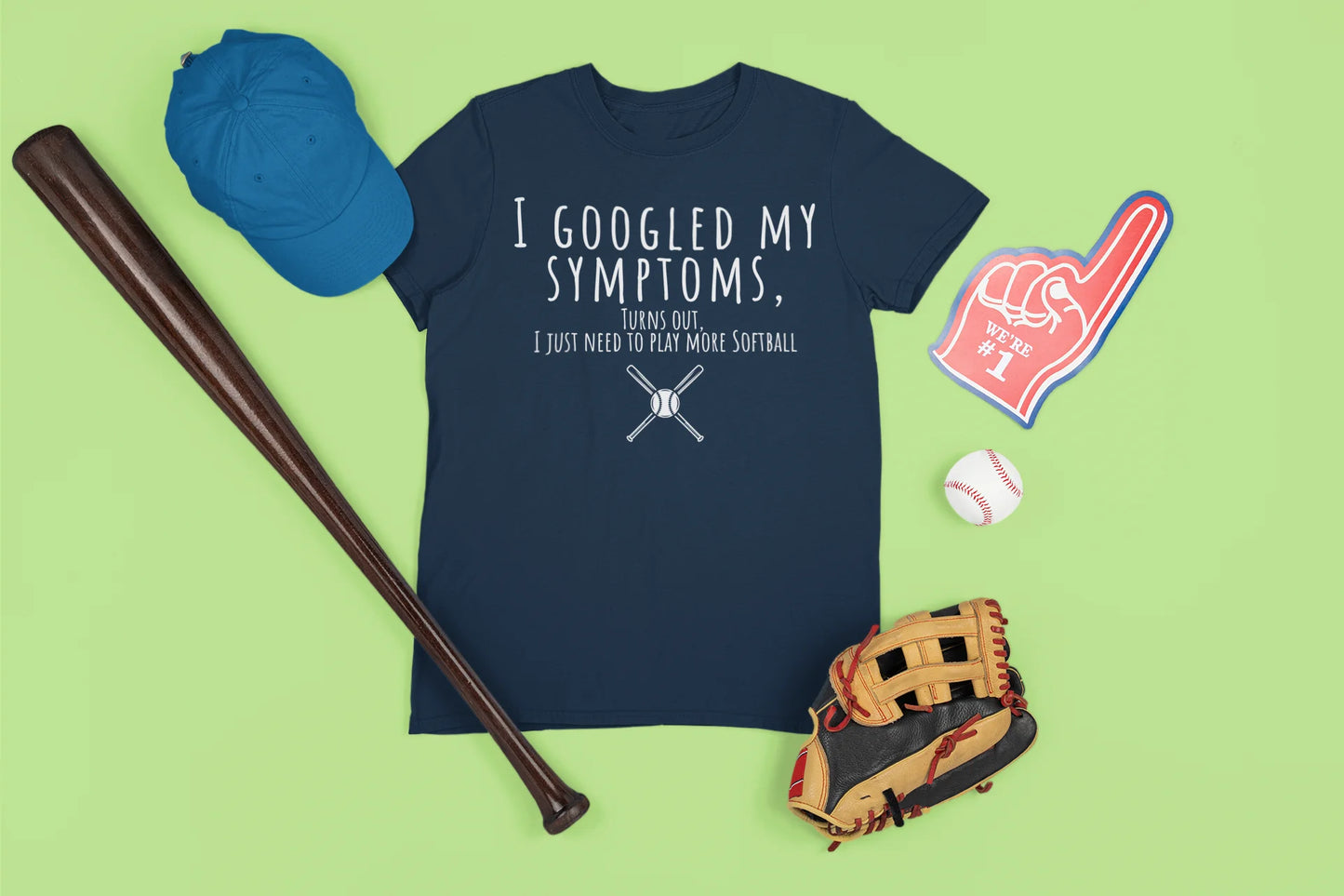 Navy t-shirt with humorous text, blue cap, baseball bat, glove, and foam finger on a green background