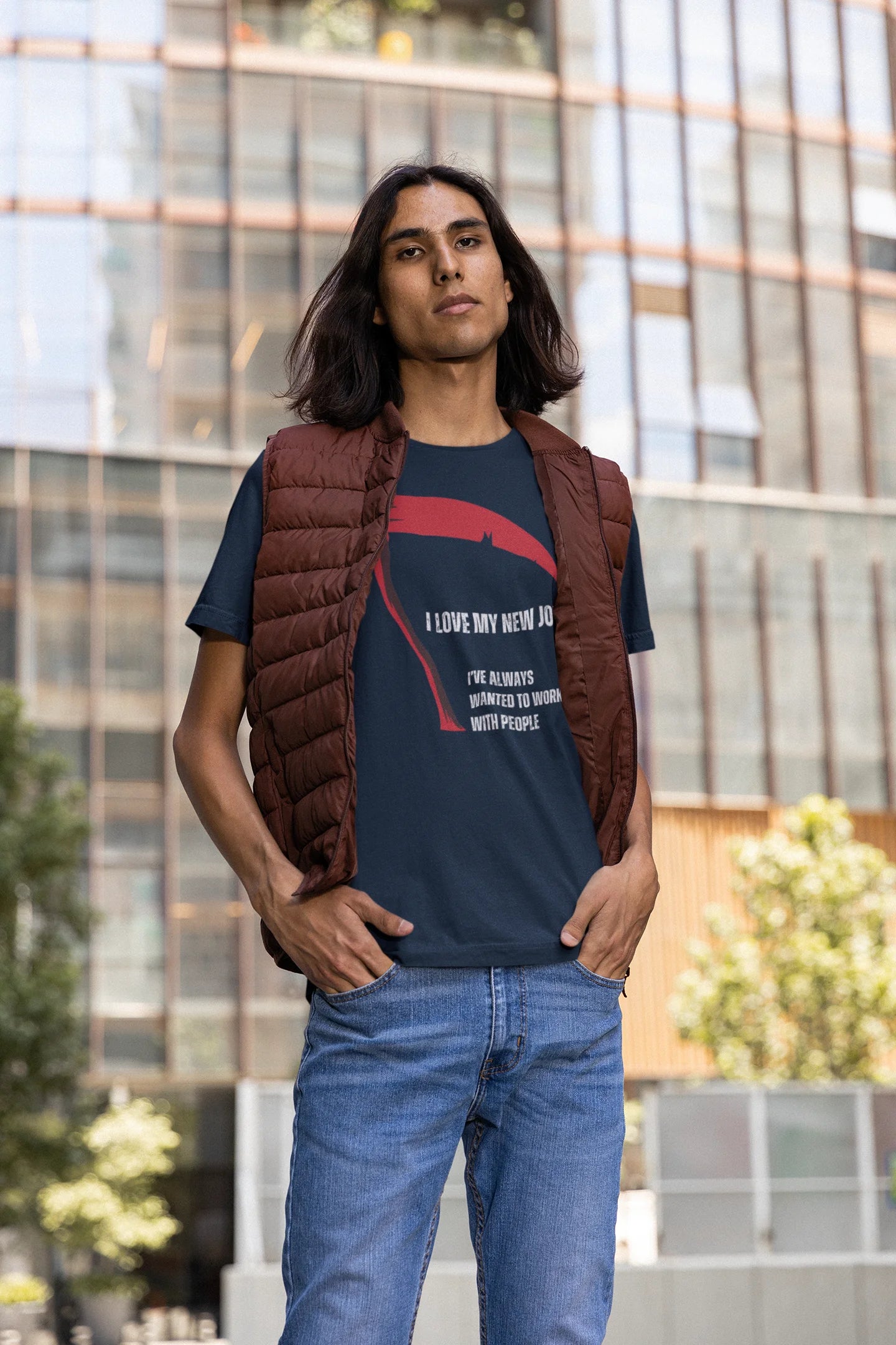 Person wearing a dark blue t-shirt with text, brown vest, and blue jeans standing in front of a modern building.