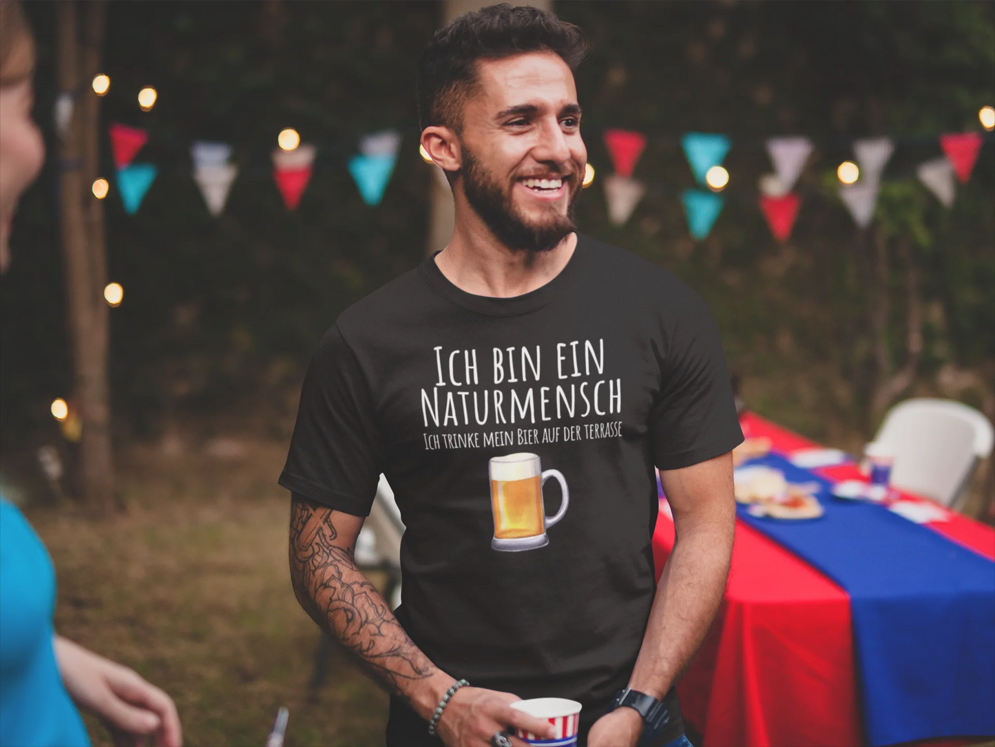 Man wearing a black t-shirt with a graphic and text, standing outdoors with a festive background.