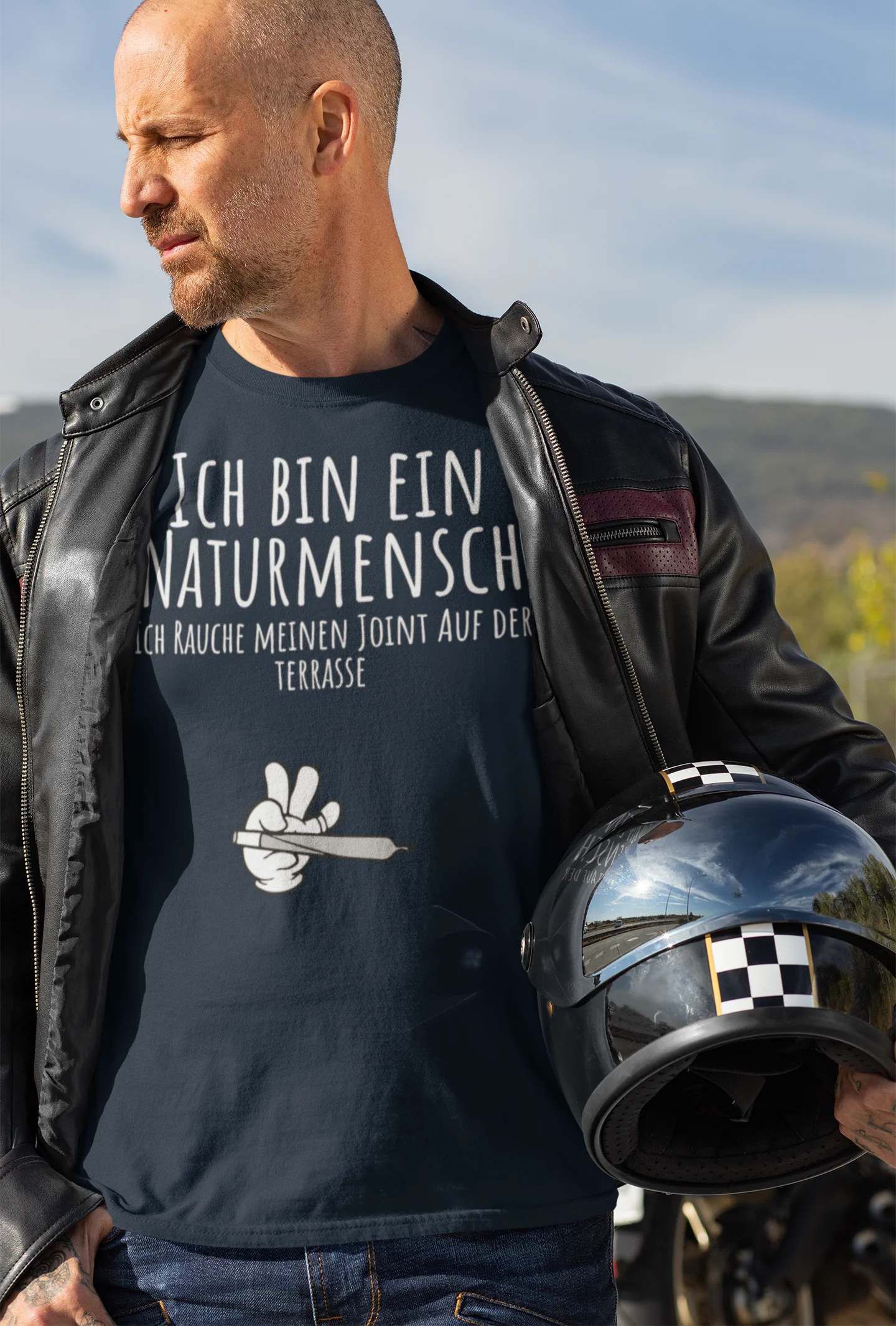 Man wearing a black t-shirt with German text and a white graphic, holding a helmet outdoors.