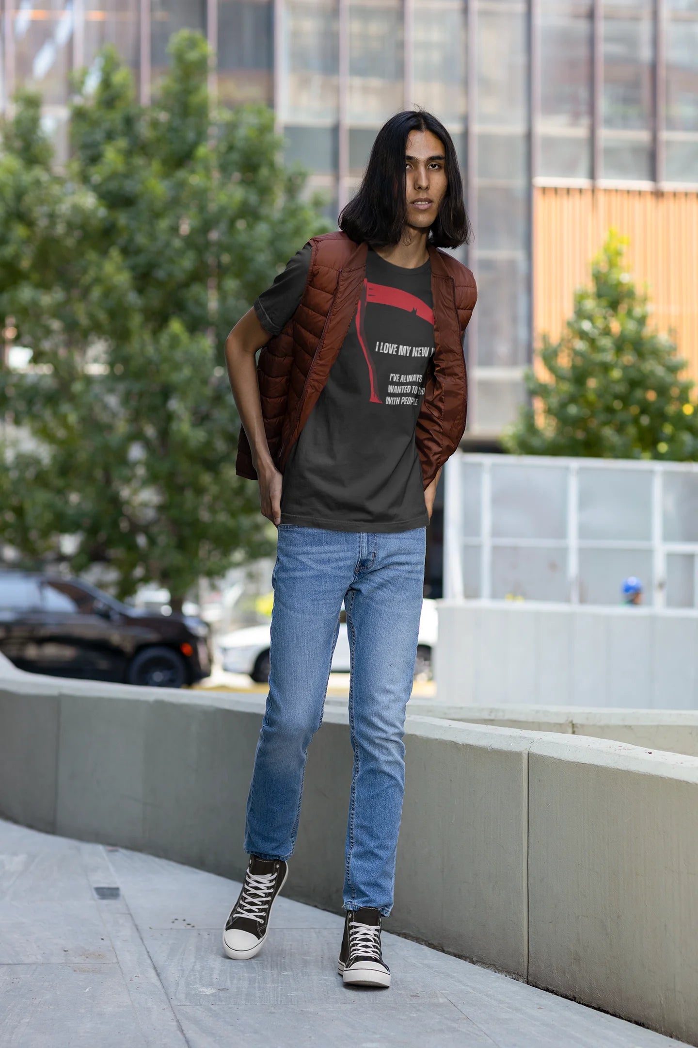 Person wearing a black t-shirt with text, blue jeans, and a brown jacket standing on a sidewalk with trees and buildings in the background.