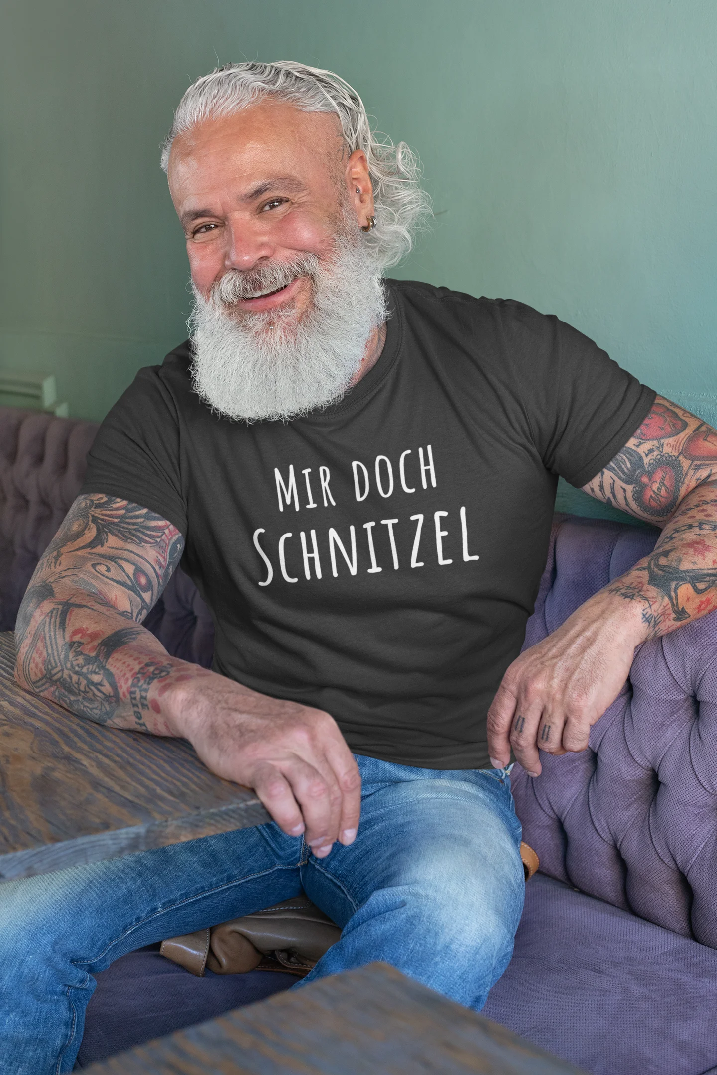 Man with tattoos and a beard wearing a black t-shirt with German text, sitting on a purple couch.