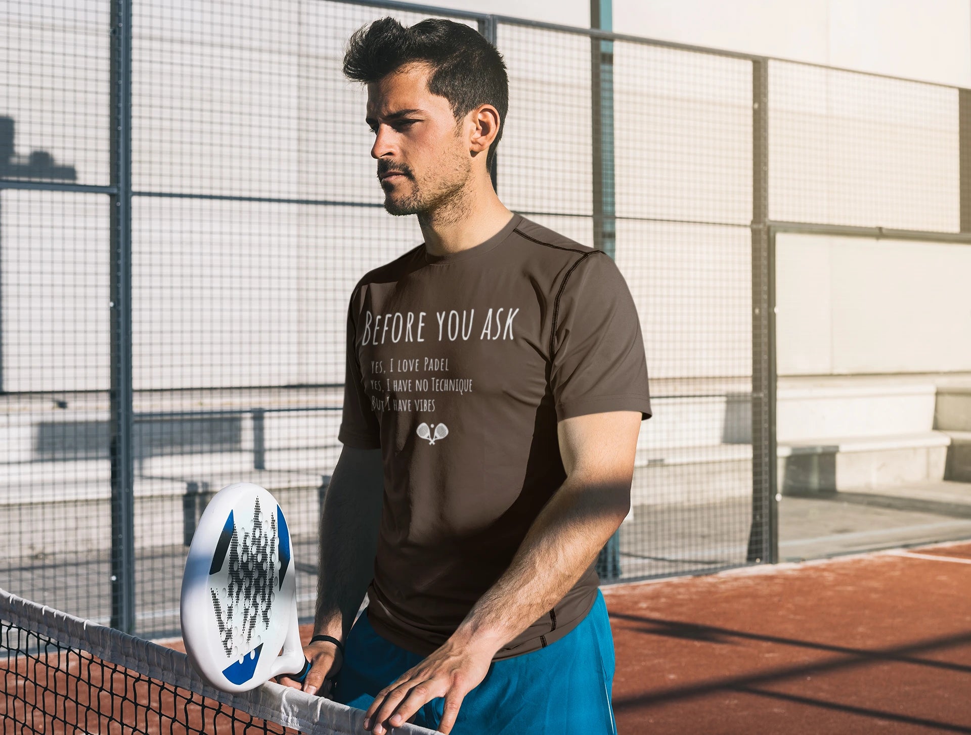 Man holding a paddle on a tennis court wearing a brown t-shirt with text.