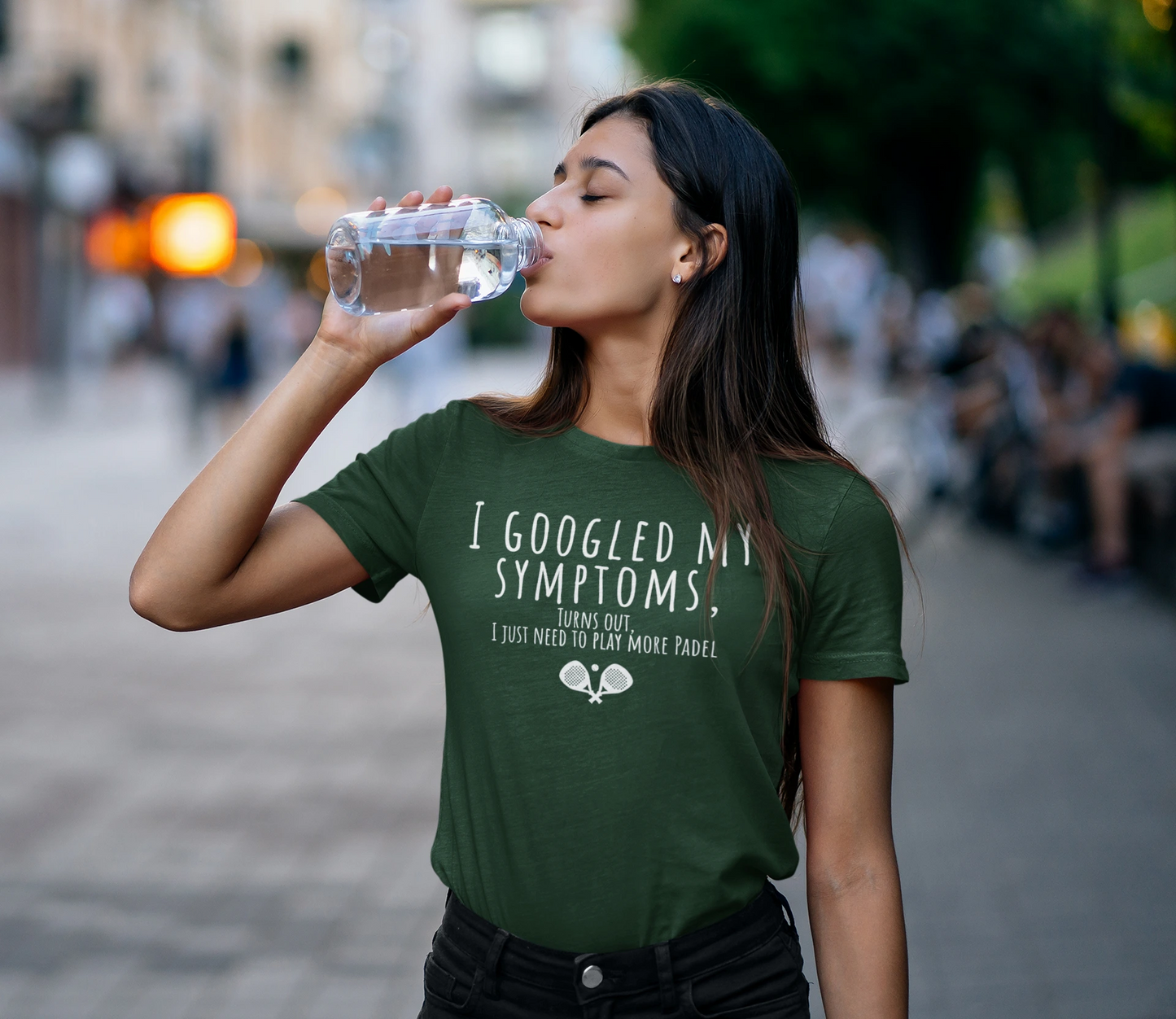 Woman drinking water outdoors wearing a green t-shirt with text.