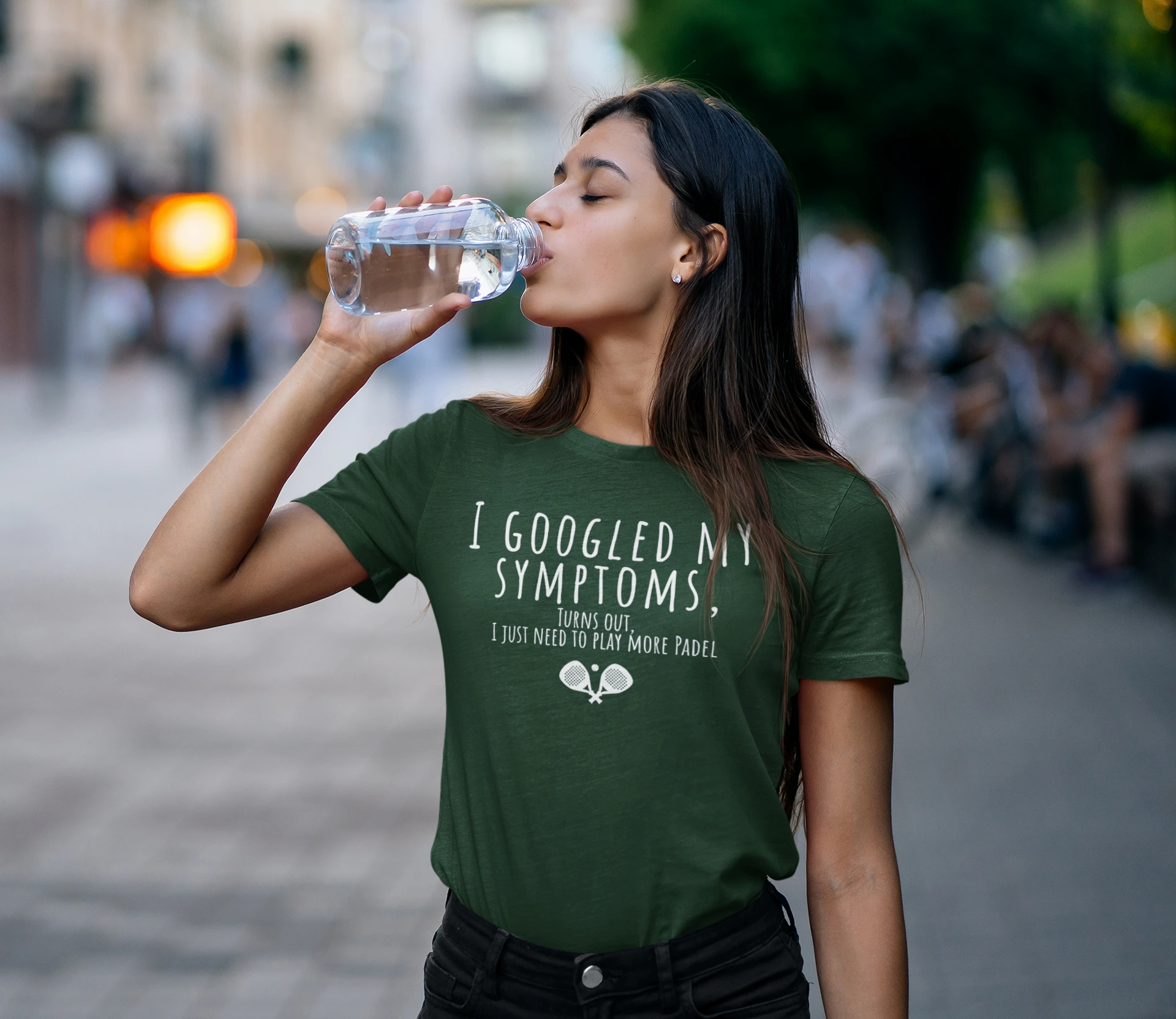 Woman drinking water outdoors wearing a green t-shirt with text.