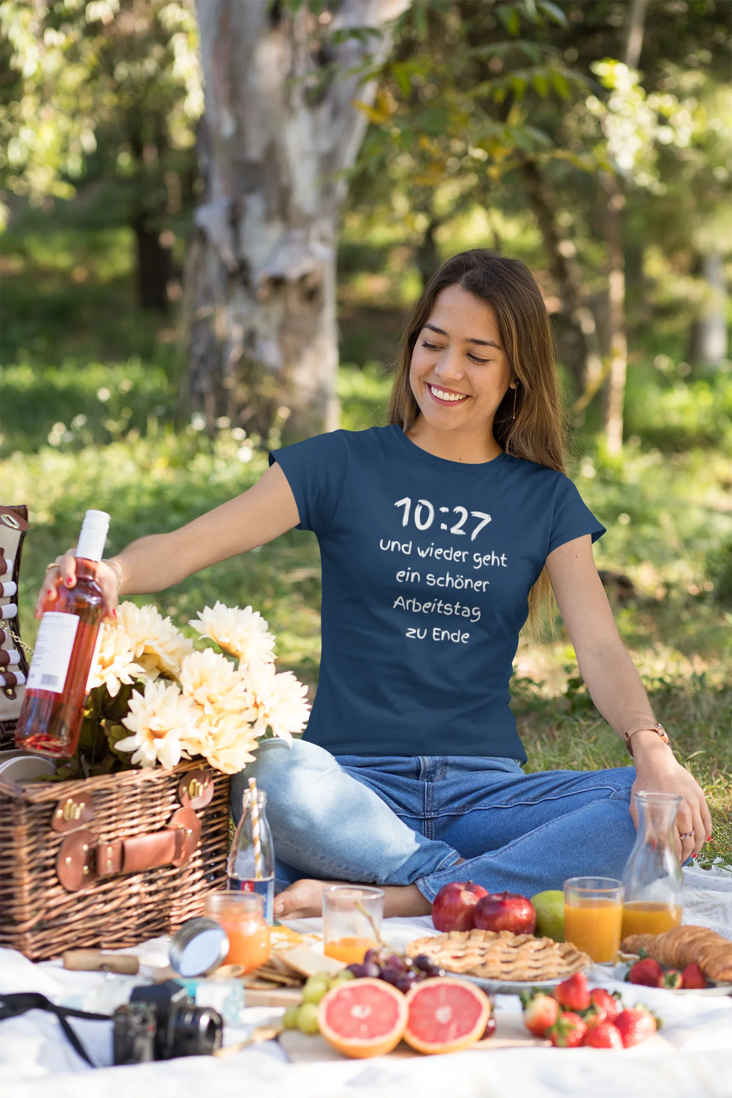 Lustiges T Shirt Woman sitting at a picnic with a blue t-shirt featuring text, surrounded by food and drinks in a natural setting.