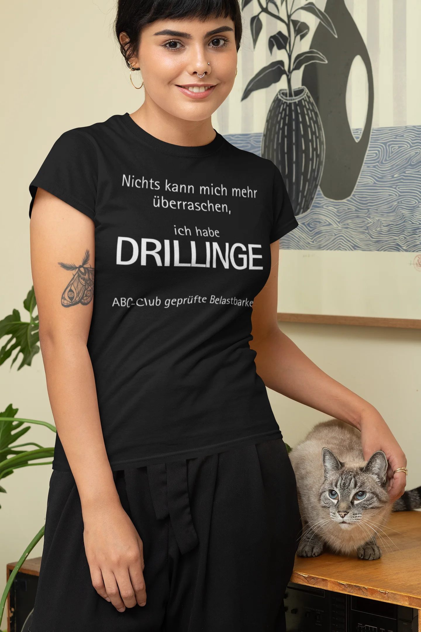 Geschenk für Drillinge Woman wearing a black t-shirt with text, sitting next to a cat indoors.
