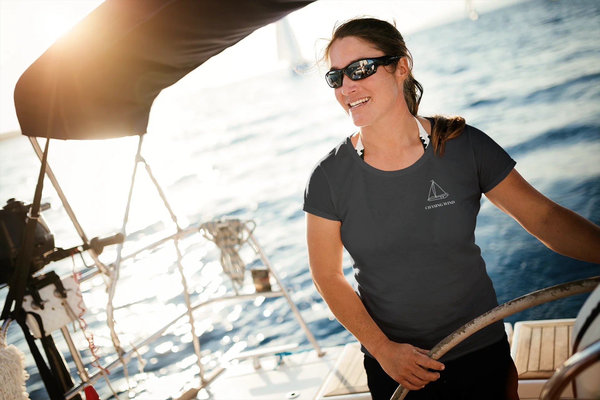 Sailing T-Shirt Woman on a sailboat wearing sunglasses and a black t-shirt with a logo, with water and sky in the background.