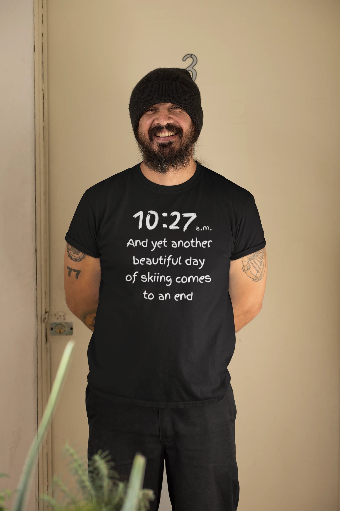 Man wearing a black t-shirt with text, standing indoors against a beige wall.