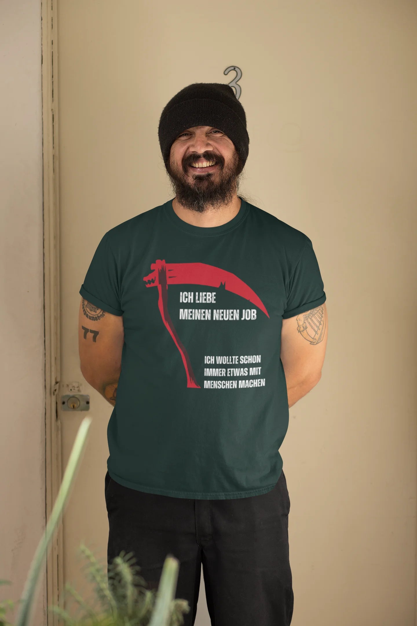 Man wearing a green t-shirt with red text and graphic, standing against a beige wall.