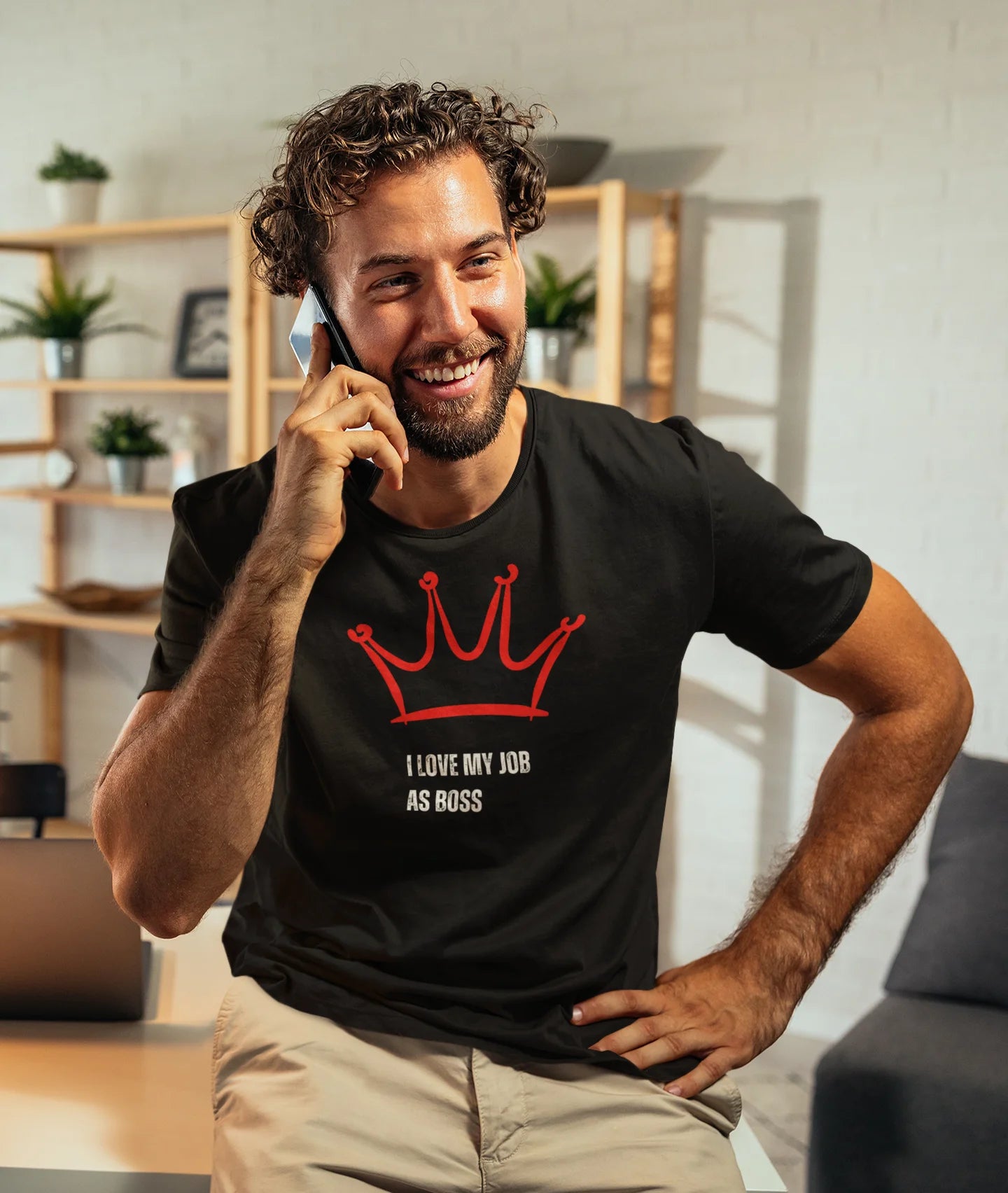 Gift for Boss Funny  Man wearing a black t-shirt with a red crown and text, talking on a phone indoors.