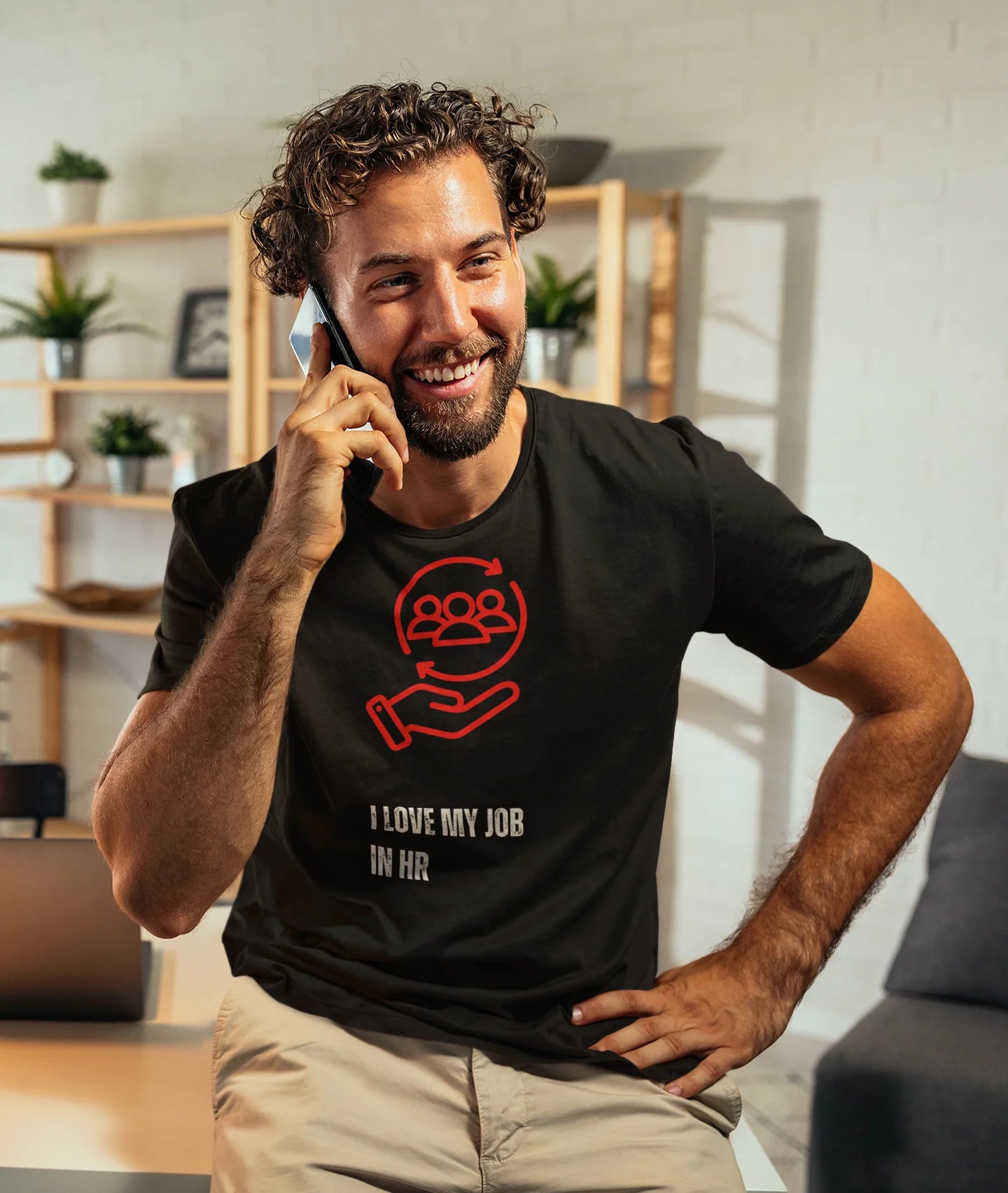 I love my Job T-Shirt Man wearing a black t-shirt with a red graphic and text, sitting in a room with shelves and plants.