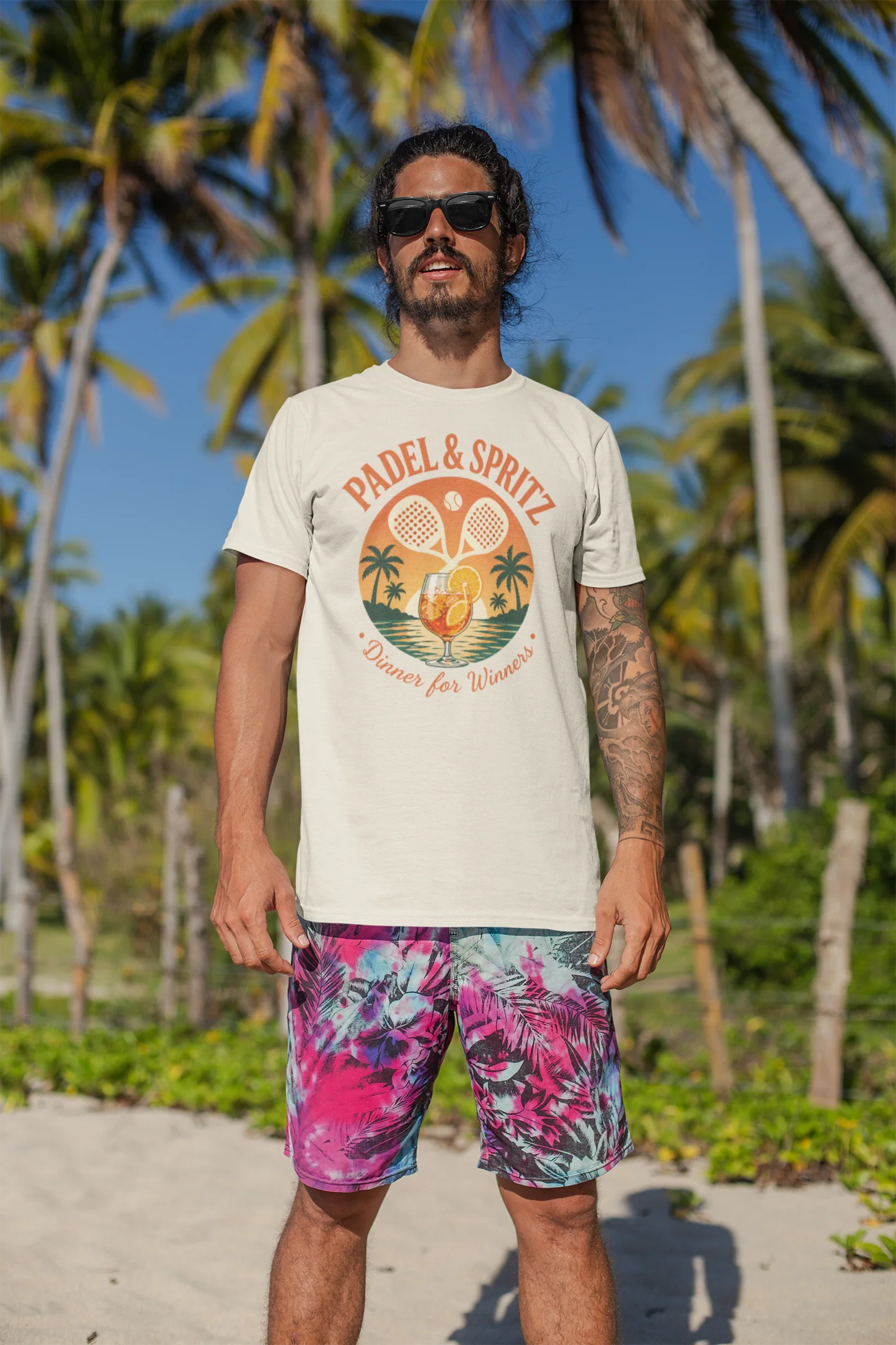 Padel Aperol T-Shirt Man wearing a 'Padel & Spritz' t-shirt and colorful shorts on a beach with palm trees.