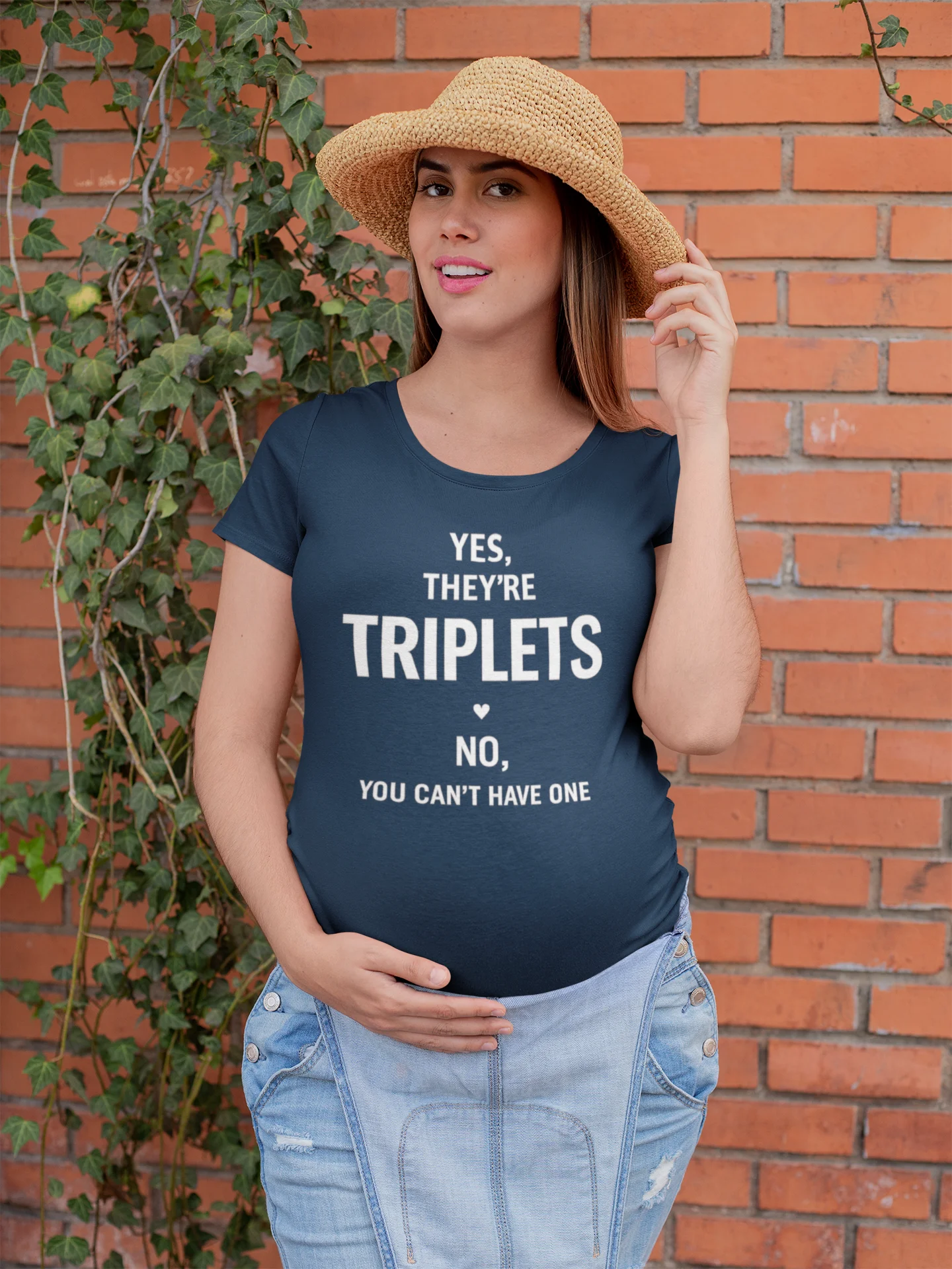 Triplets Mom T-Shirt Woman wearing a navy blue t-shirt with humorous text, standing against a brick wall.