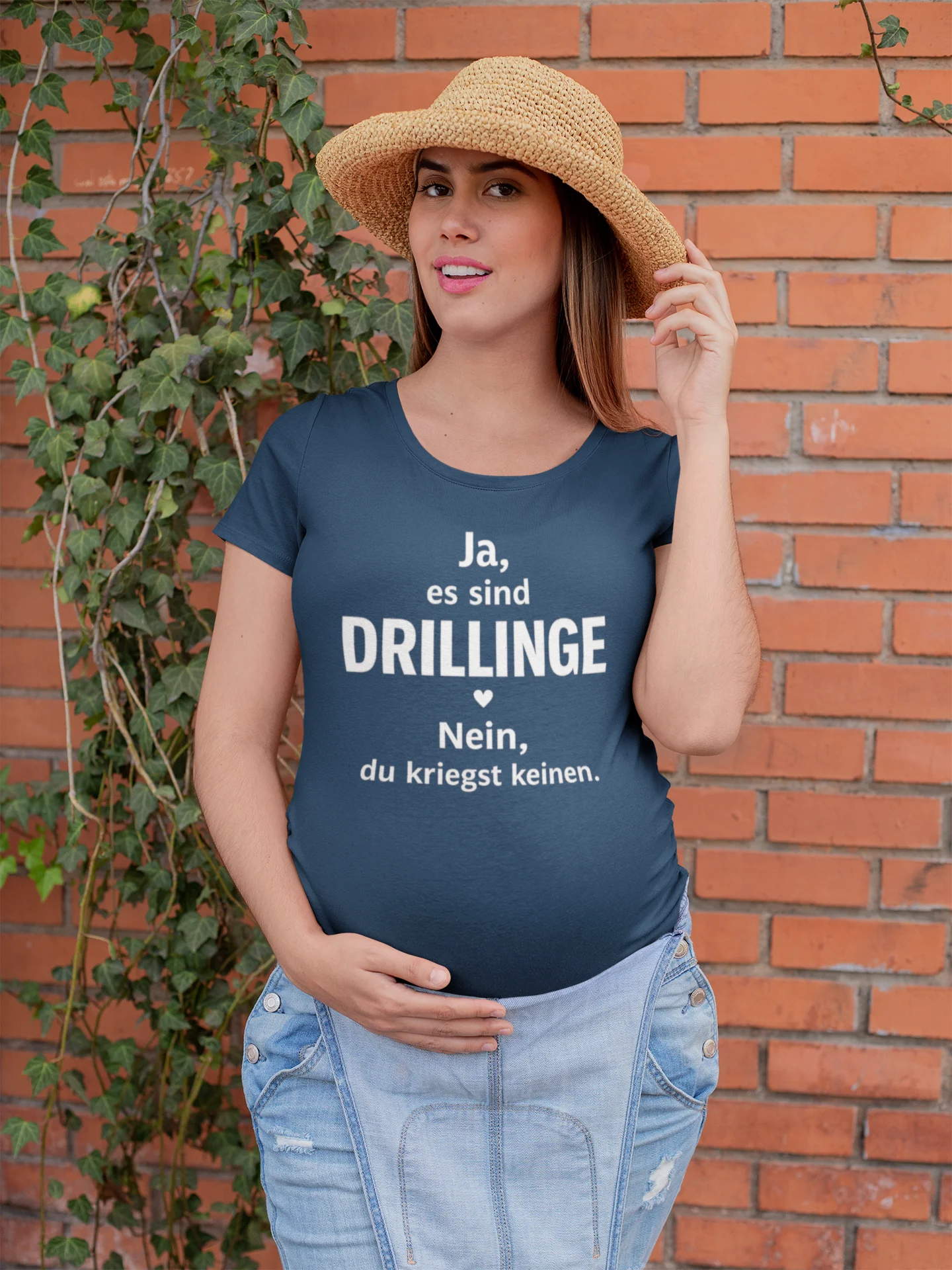 T-Shirt für Drillingseltern Woman wearing a navy blue t-shirt with German text against a brick wall.
