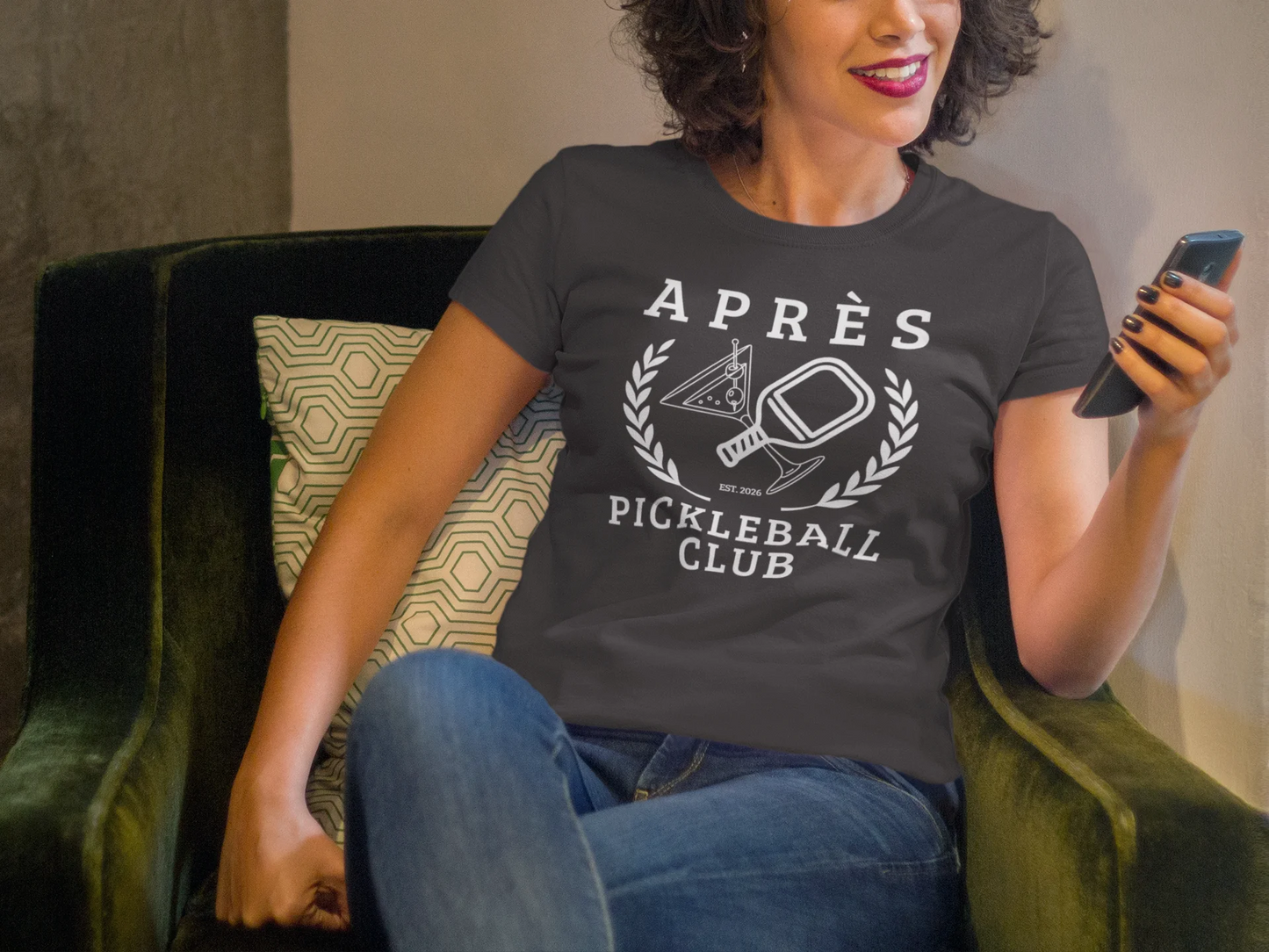 Funny Pickleball Shirt Woman wearing a t-shirt with 'Après Pickleball Club' design, sitting on a green chair.