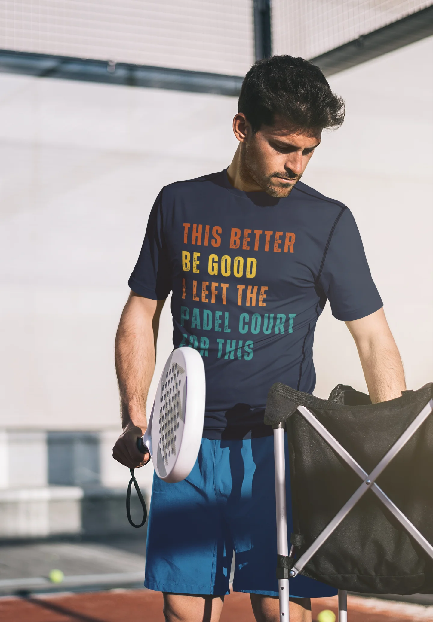 Man holding a racquet and a bag, wearing a t-shirt with a humorous message, on a padel court.